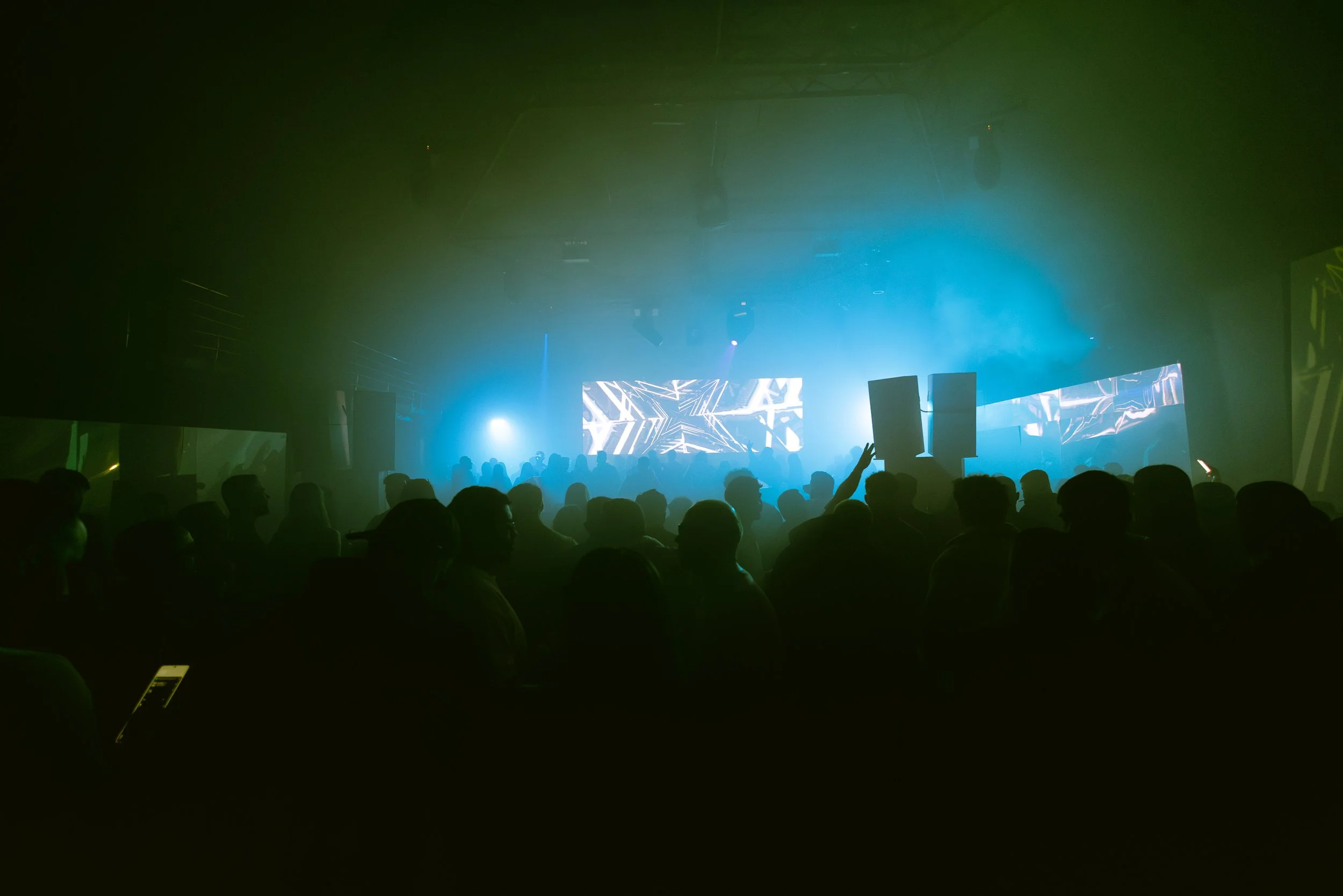 Crowd of people dancing and enjoying a music concert with bright blue and white lights and digital screens displaying abstract patterns.