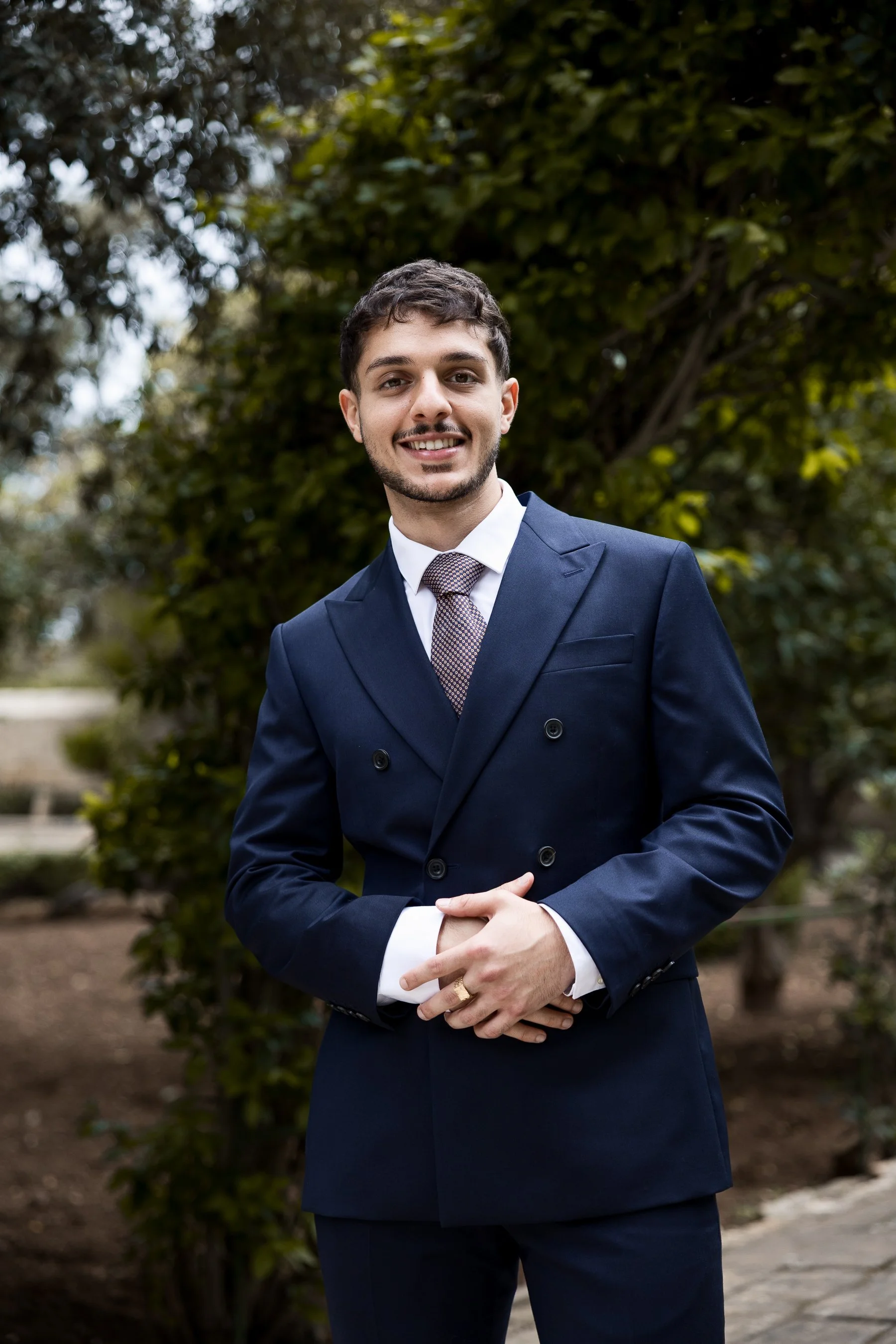 A young man with dark brown hair, a beard, wearing a navy blue double-breasted suit, white dress shirt, and patterned tie, standing outdoors in front of green foliage, smiling at the camera.