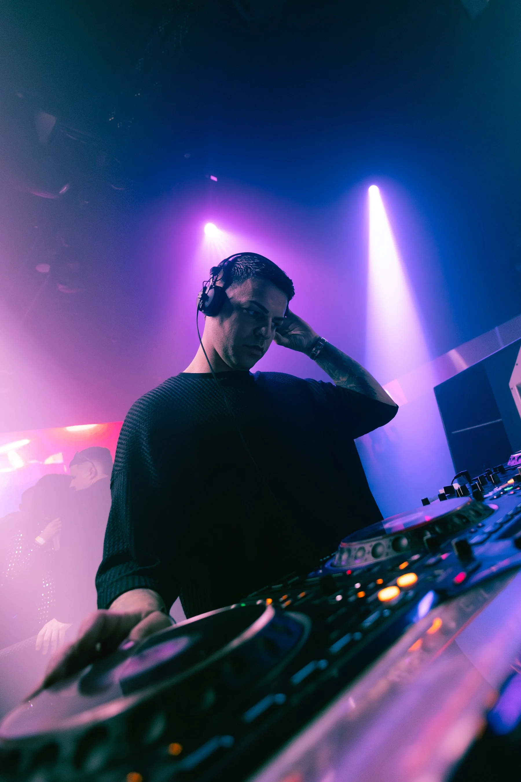 A DJ wearing headphones adjusts equipment in a nightclub with purple and pink lighting.