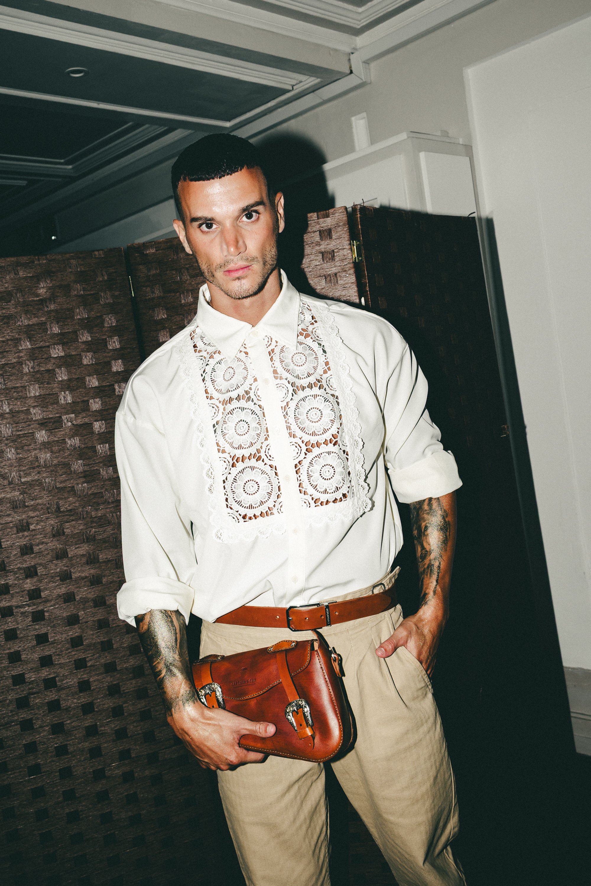 Man wearing a cream-colored shirt with lace detailing, beige pants, and holding a brown leather bag, standing indoors with a woven partition in the background.