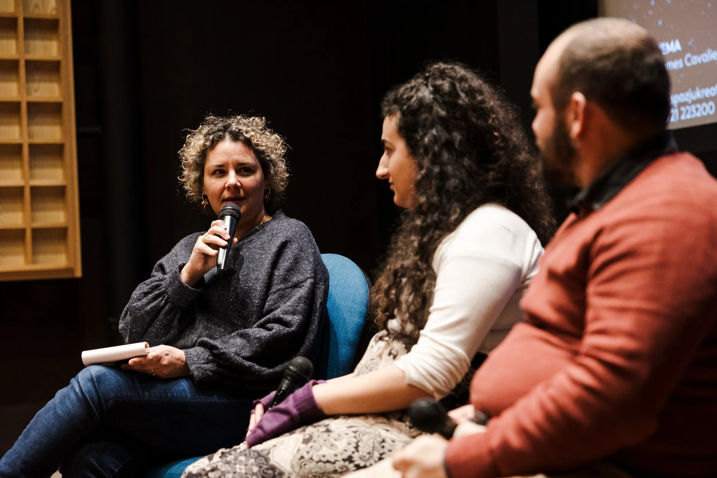 A woman with curly hair holding a microphone and a notepad, speaking at a panel discussion with two other people, a woman and a man, seated beside her on a stage.