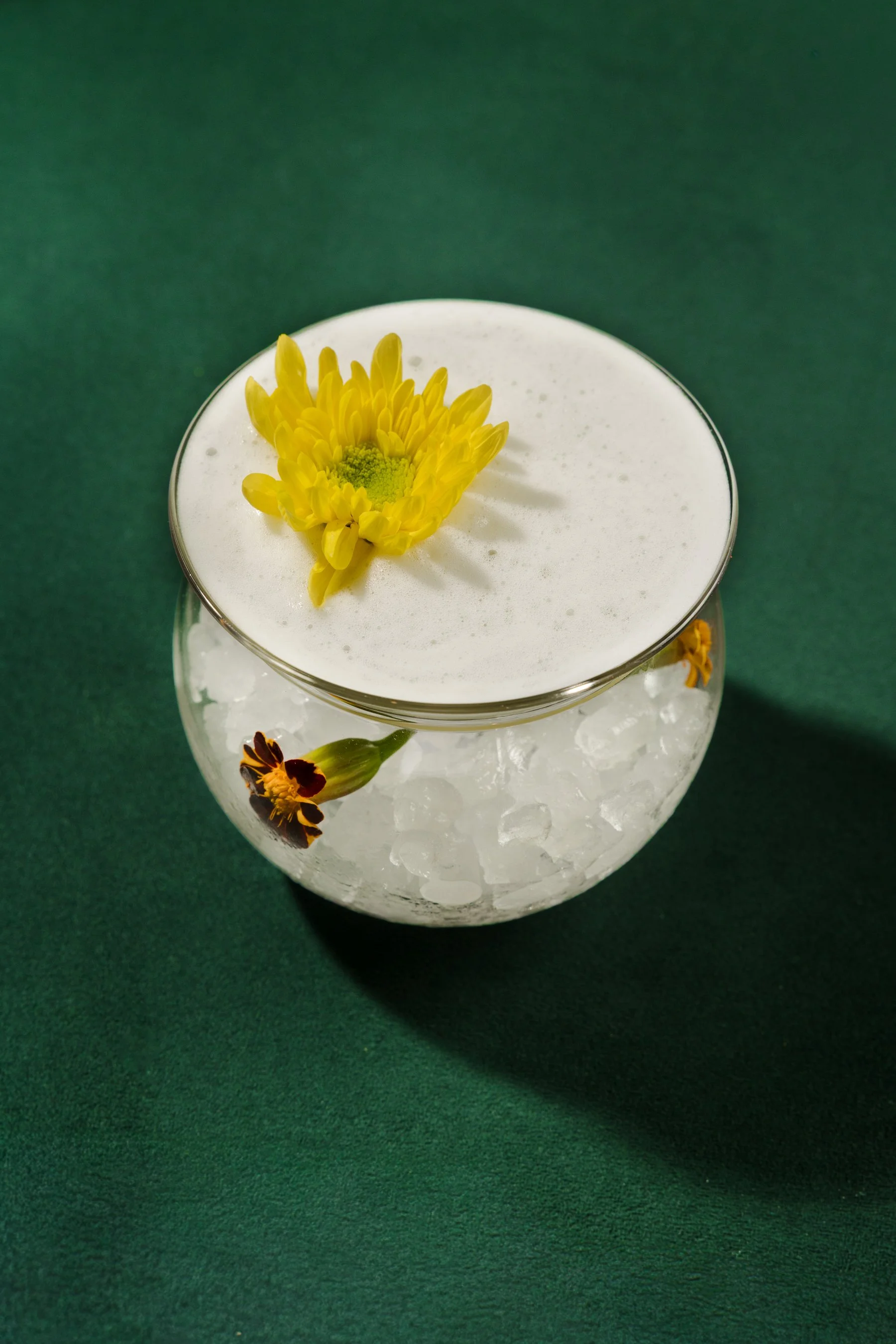 A glass filled with ice cubes and topped with a foam layer, decorated with a yellow flower and a small dark flower petal on a green background.