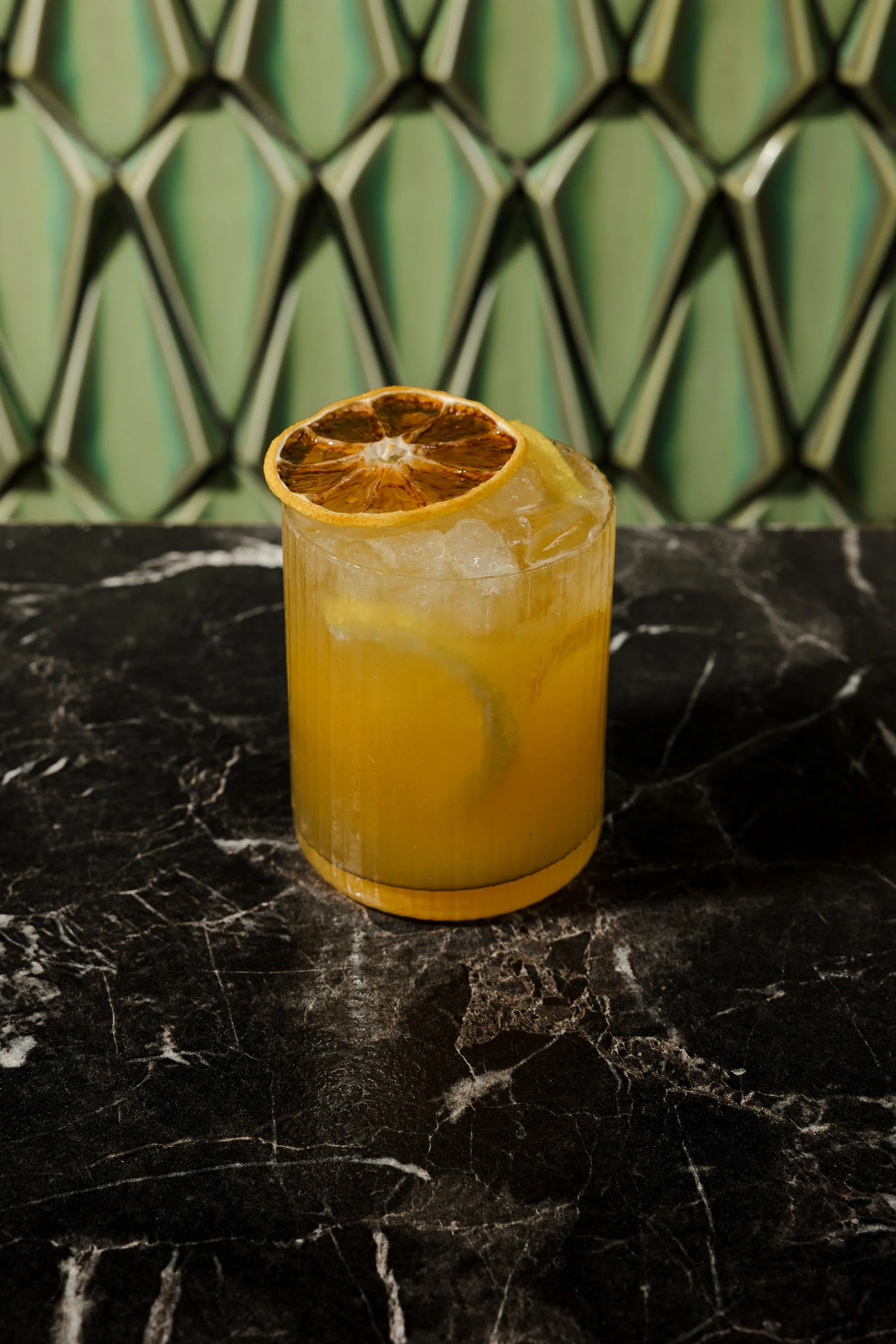 Yellow drink with ice in a tall glass, garnished with a dried lemon slice, on a black marble surface with a patterned green background.