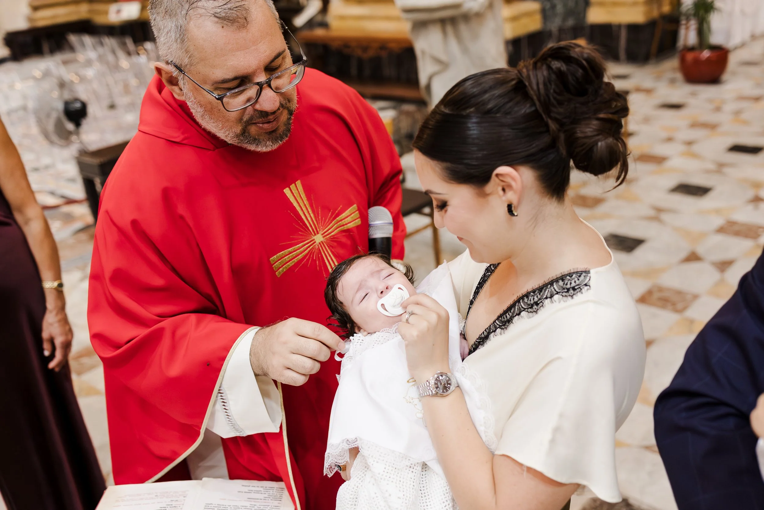 A woman holding a baby during a baptism ceremony, with a priest dressed in red performing the ritual.