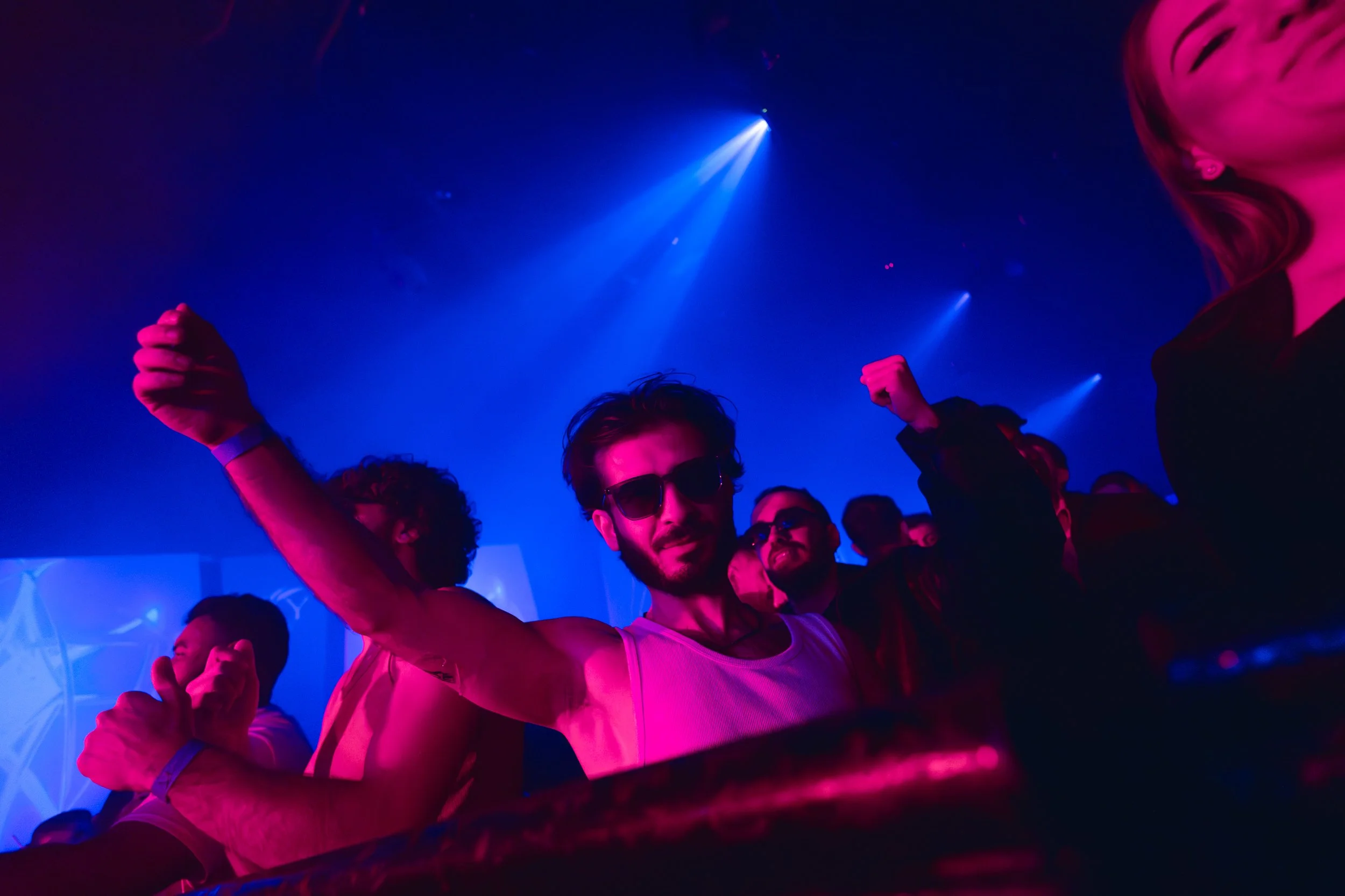 People dancing and enjoying at a nightclub with vibrant pink and blue lighting, some wearing sunglasses, celebrating and raising their fists in the air.