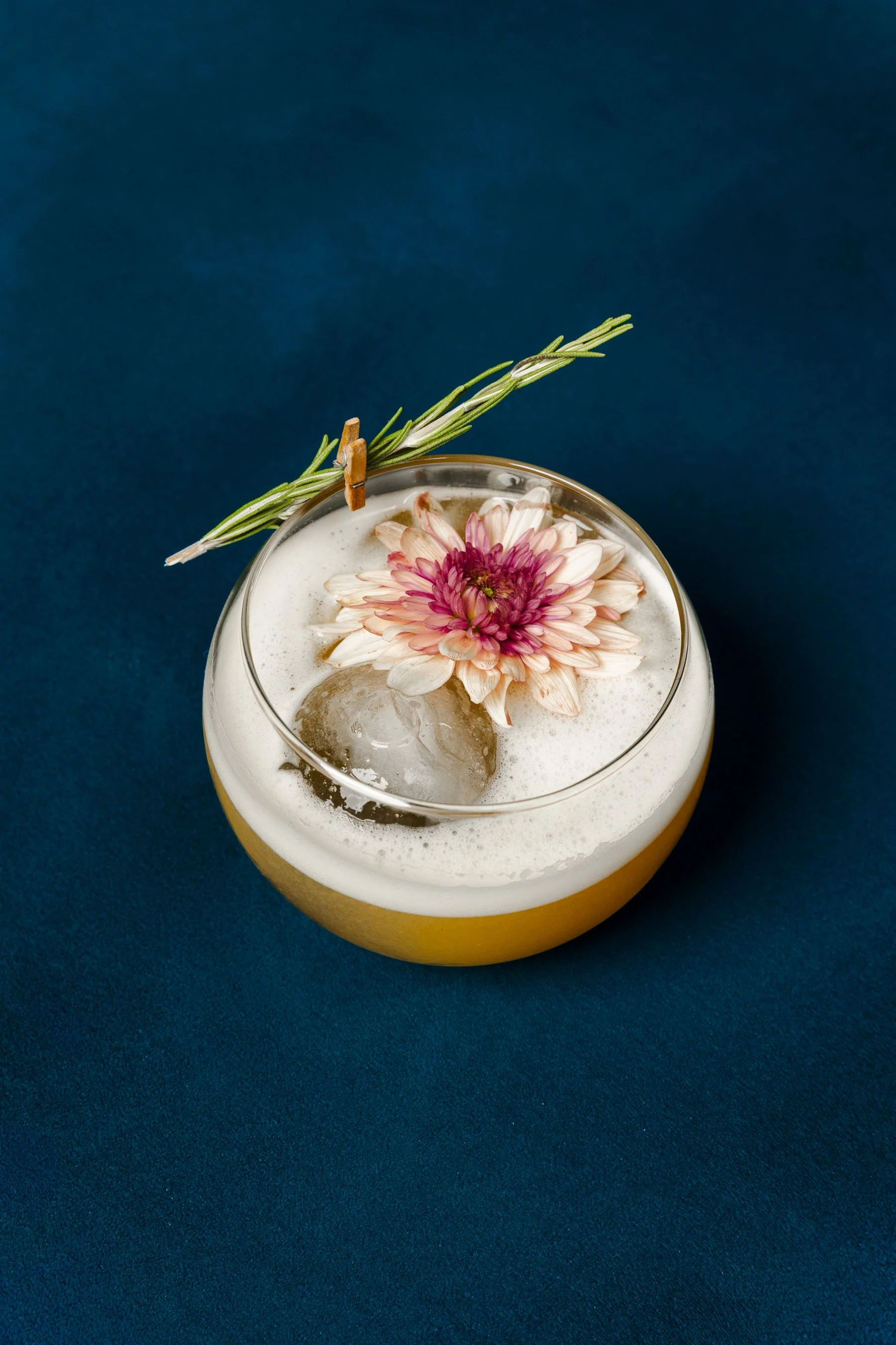 A glass of a yellow effervescent drink garnished with a pink chrysanthemum flower and a sprig of rosemary, set against a dark blue background.