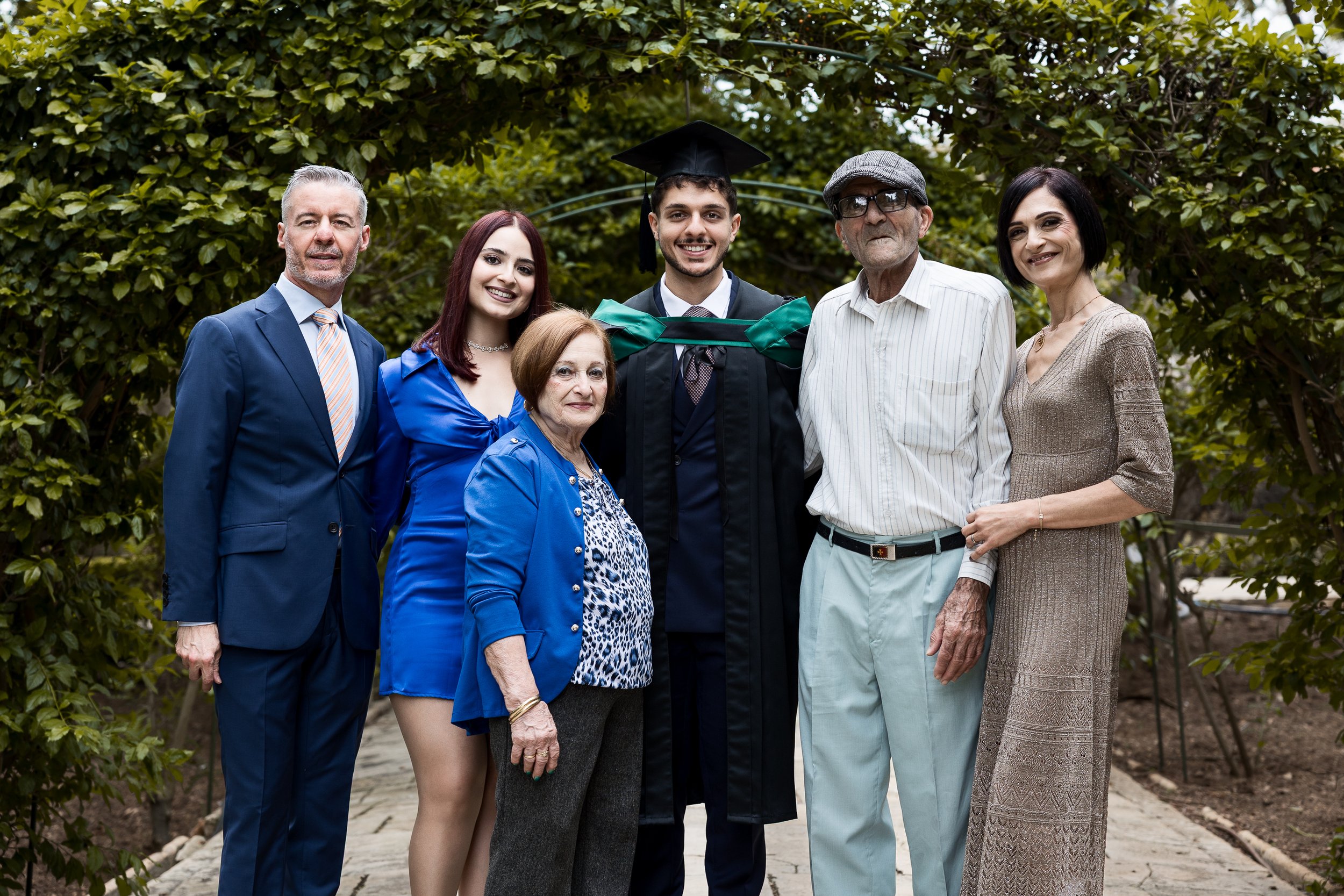 Group of seven people celebrating graduation outdoors, including a young man in cap and gown surrounded by family members.