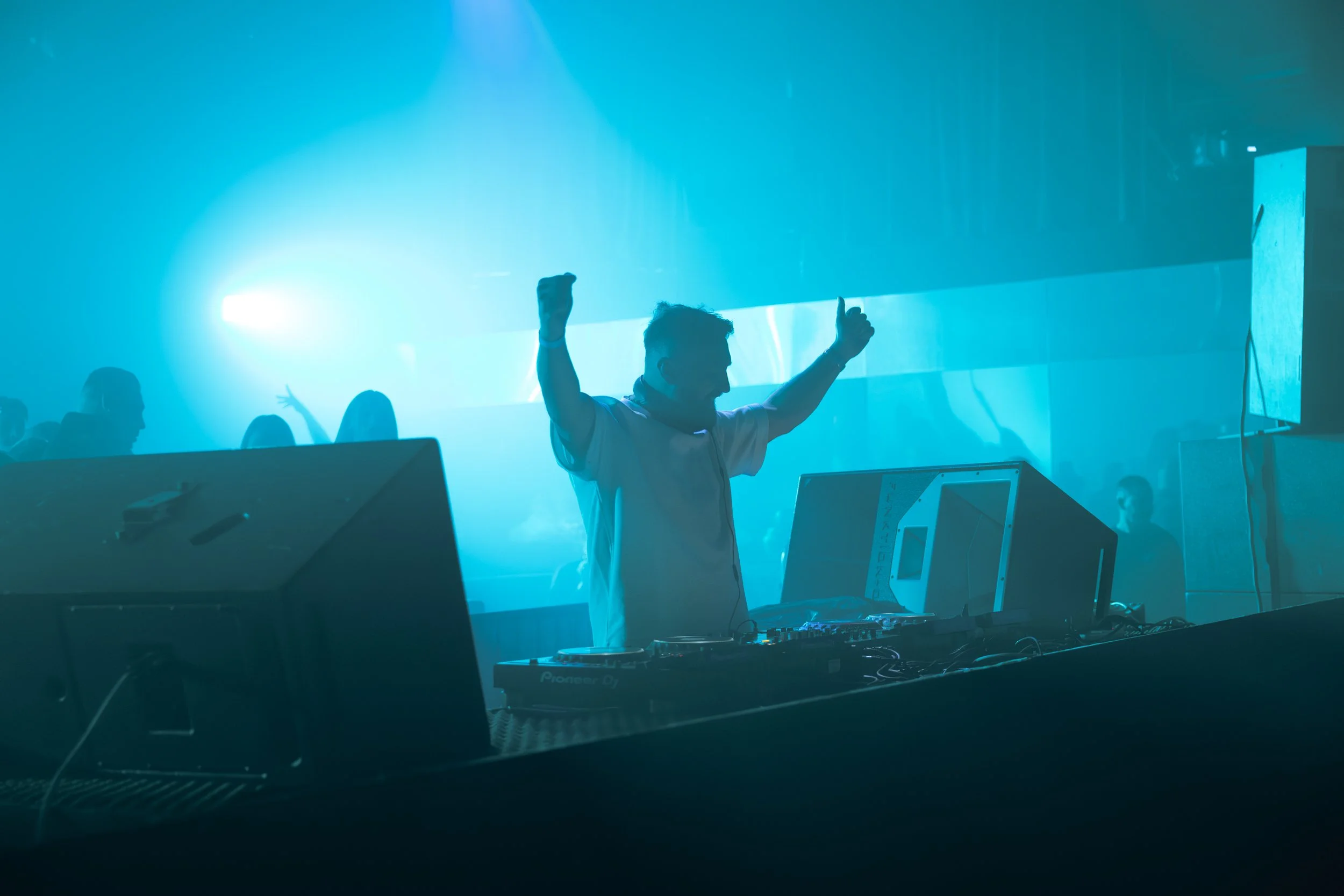A DJ is performing at a nightclub with blue lighting, wearing a white shirt, with hands raised and finger pointing up, while people dance in the background.