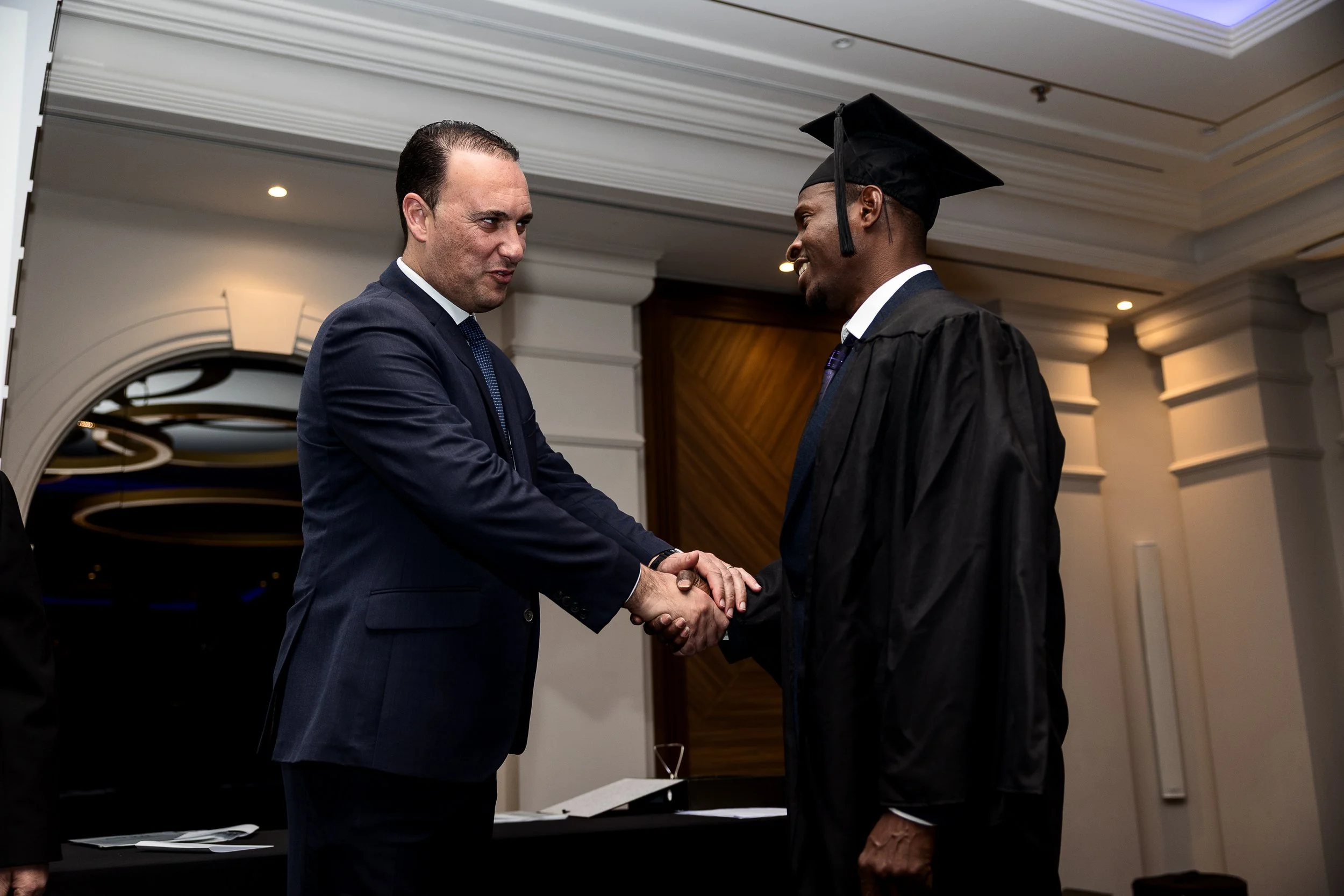 A man in a suit shaking hands with a man in a graduation gown and cap at an indoor ceremony.