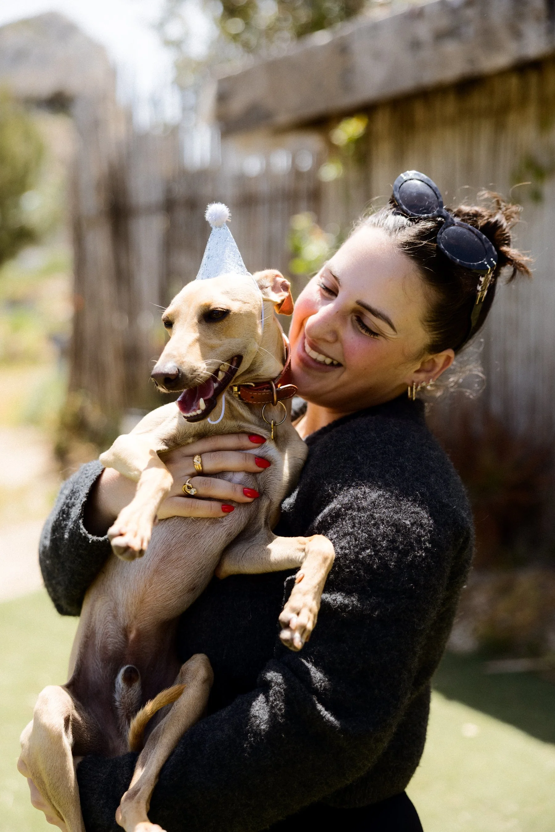 A woman smiling and holding a tan dog wearing a small white party hat outdoors. The woman has sunglasses on her head and is wearing a black and gray sweater.