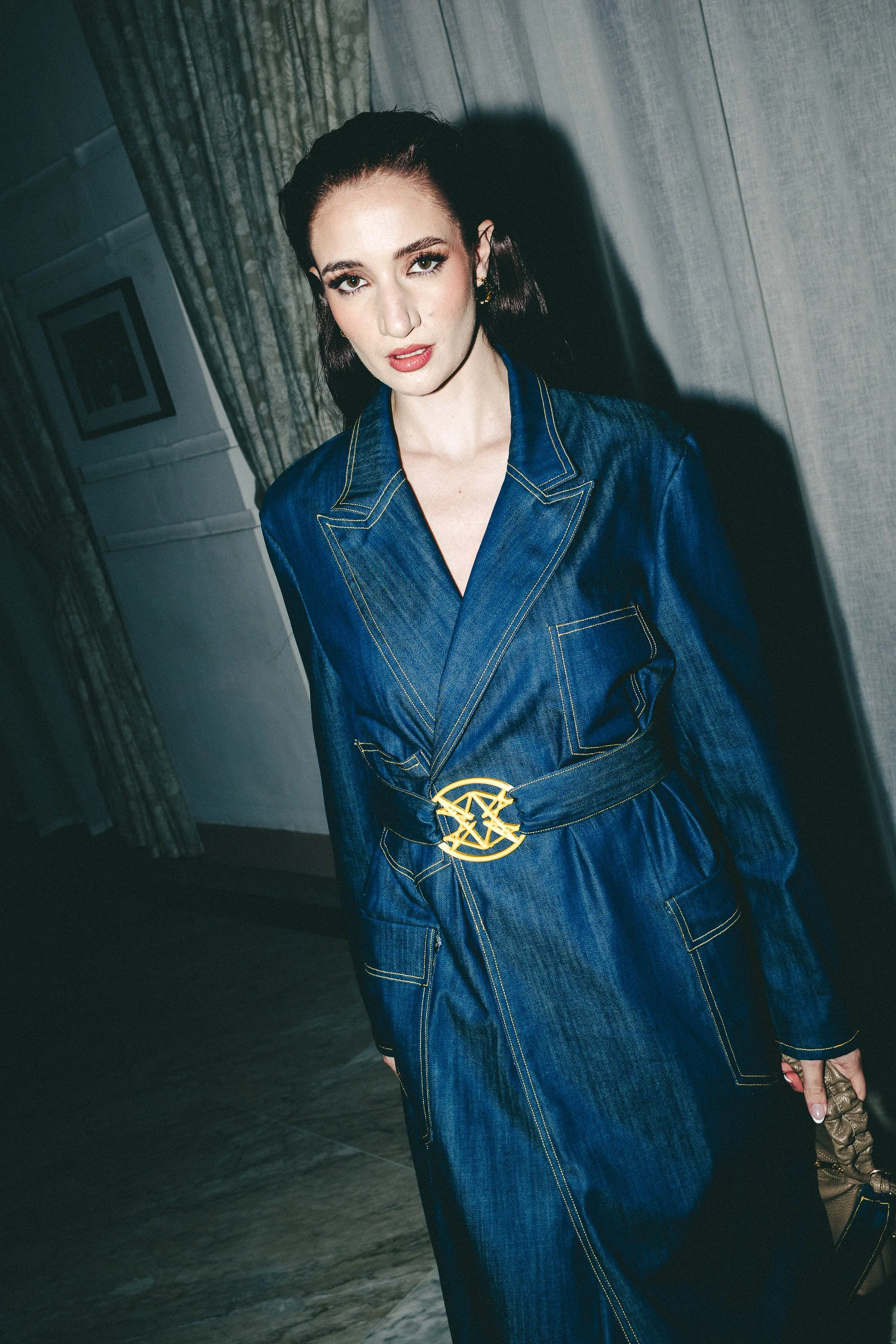 A woman in a denim jumpsuit with gold stitching and a gold belt buckle, standing inside a room with curtains and wall art.