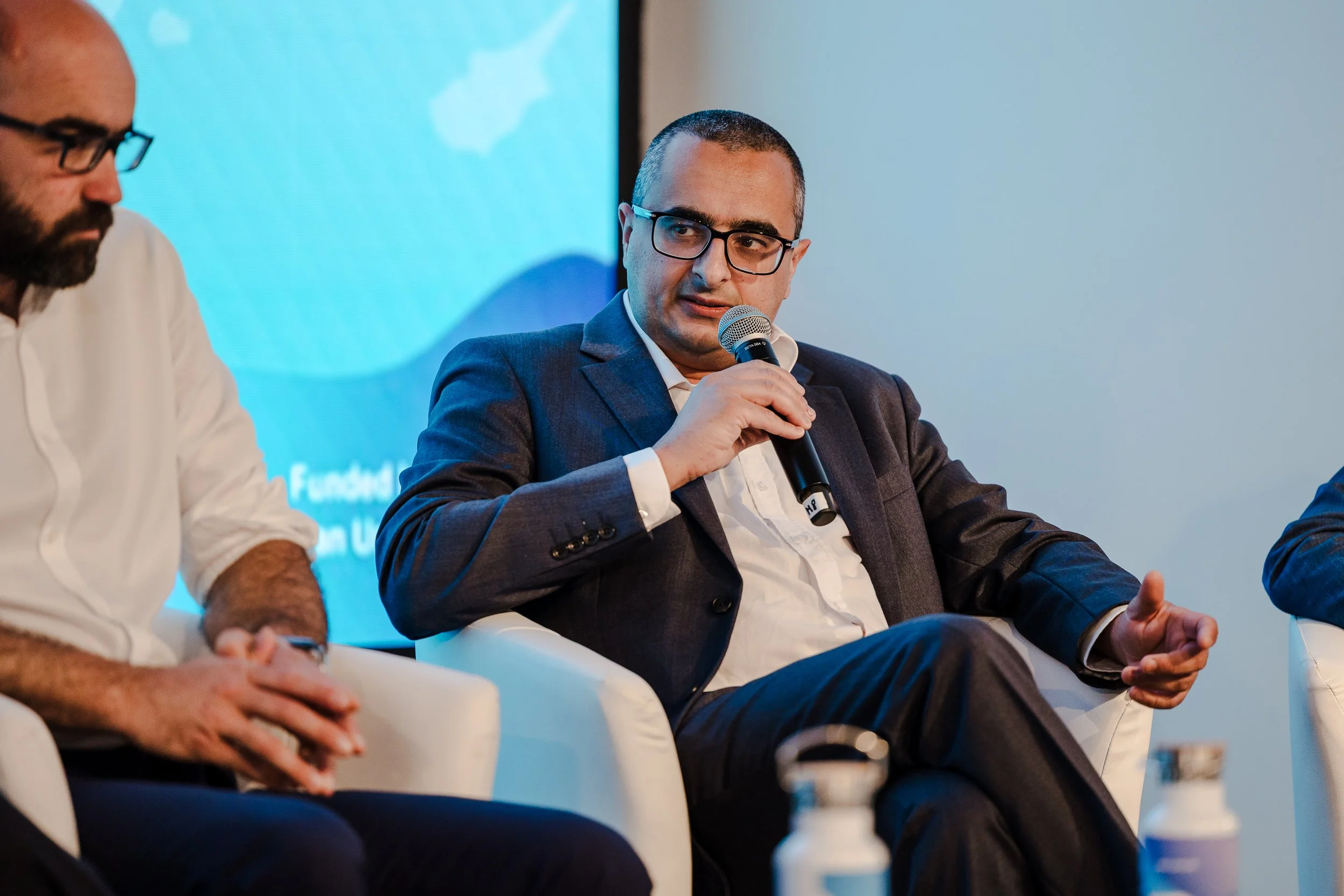Man in a dark suit and glasses speaking into a microphone during a panel discussion, seated on a white chair.
