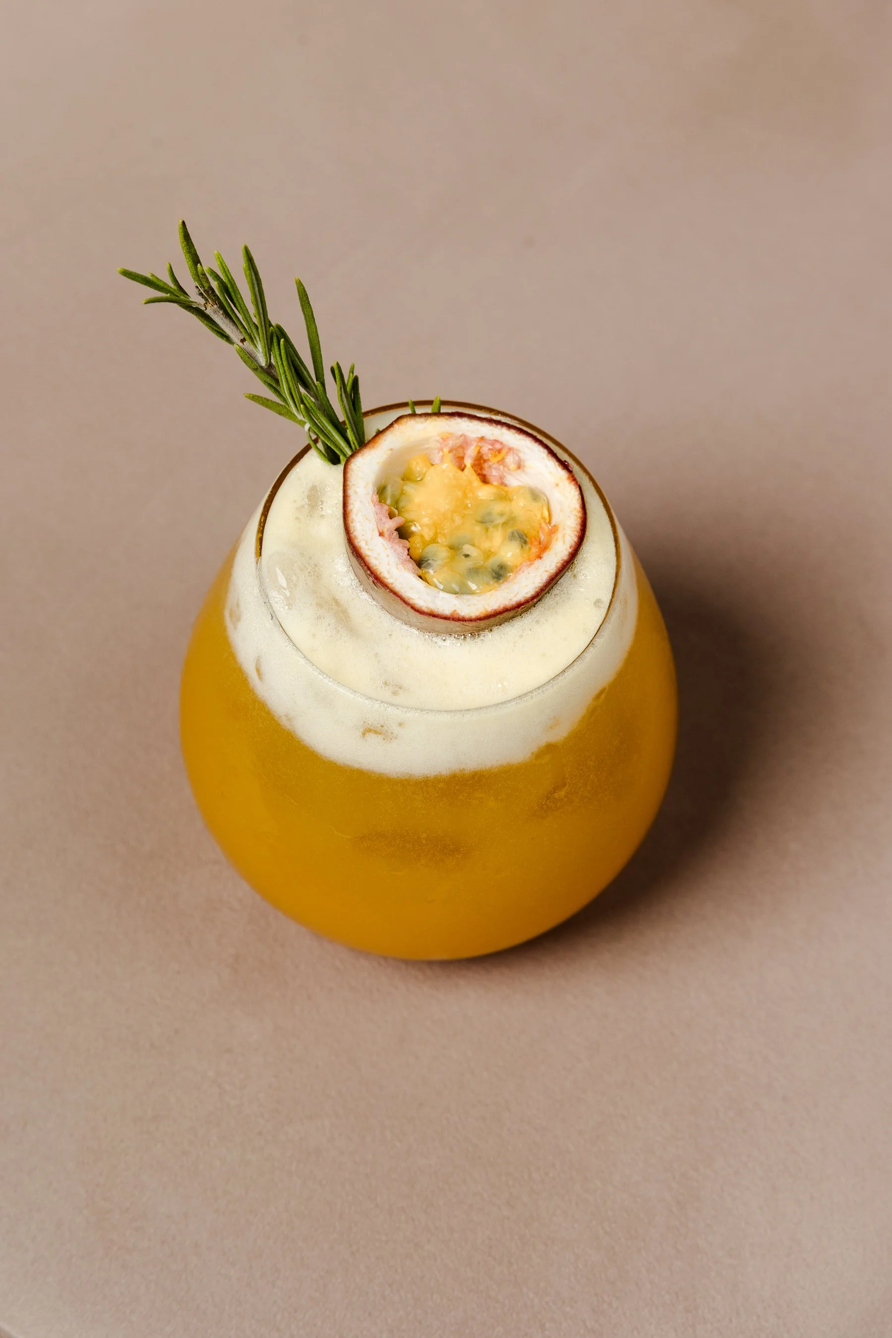 A yellow pear-shaped glass filled with a yellowish beverage, topped with a halved passion fruit and a sprig of rosemary.