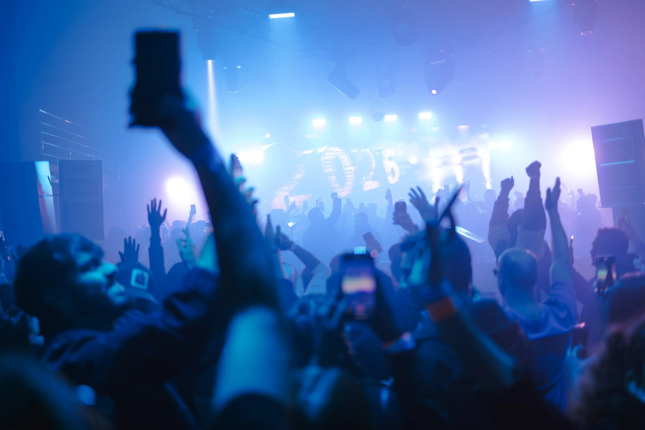 Concert crowd with raised hands and smartphones, stage with bright blue and purple lights, and large digital display showing '2026' in the background.