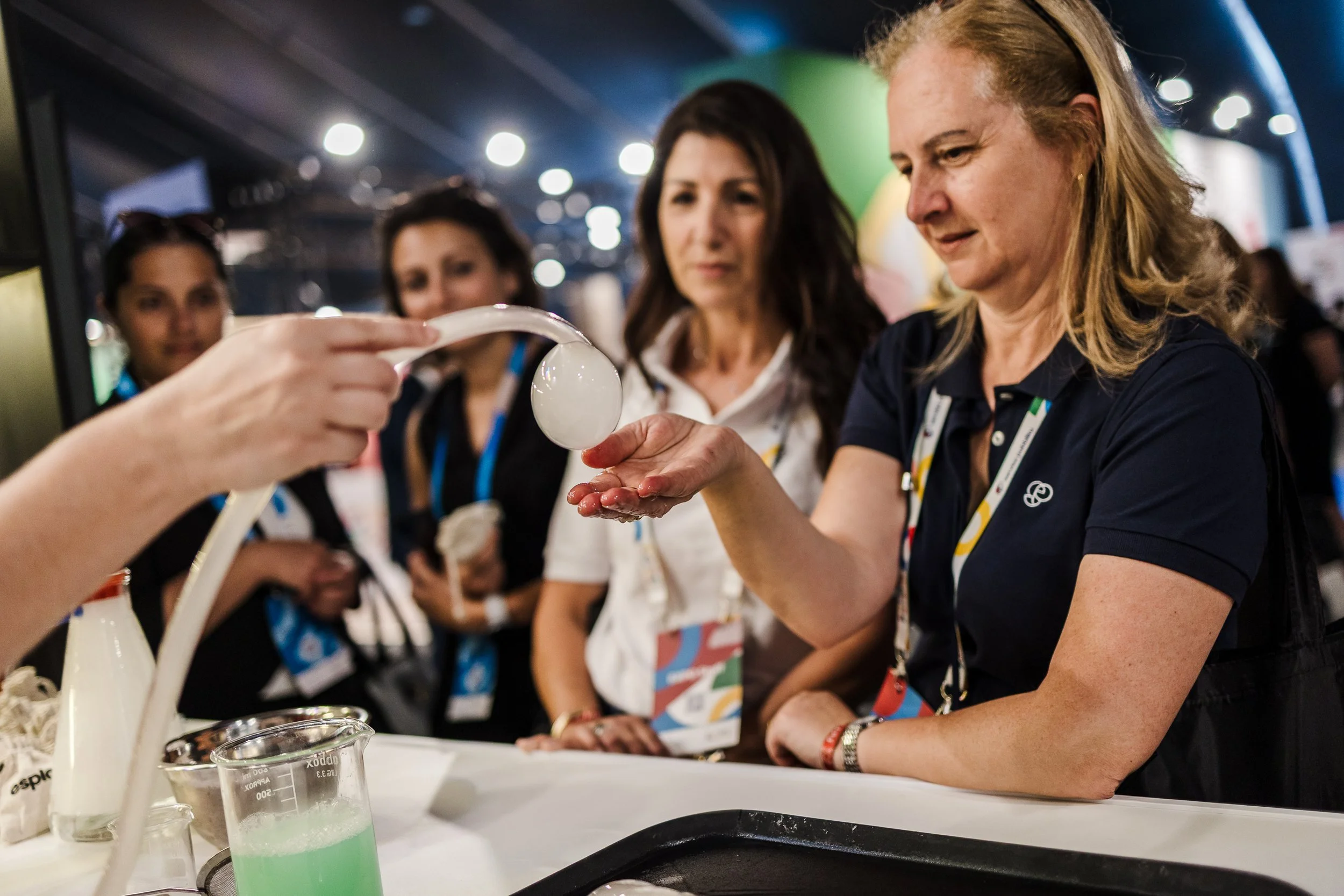 A woman is demonstrating the use of a liquid nitrogen ice cream maker to a group of women at an event. One woman is holding her hand out as the woman in the foreground pours liquid nitrogen into her hand from a spoon. The onlookers are observing with