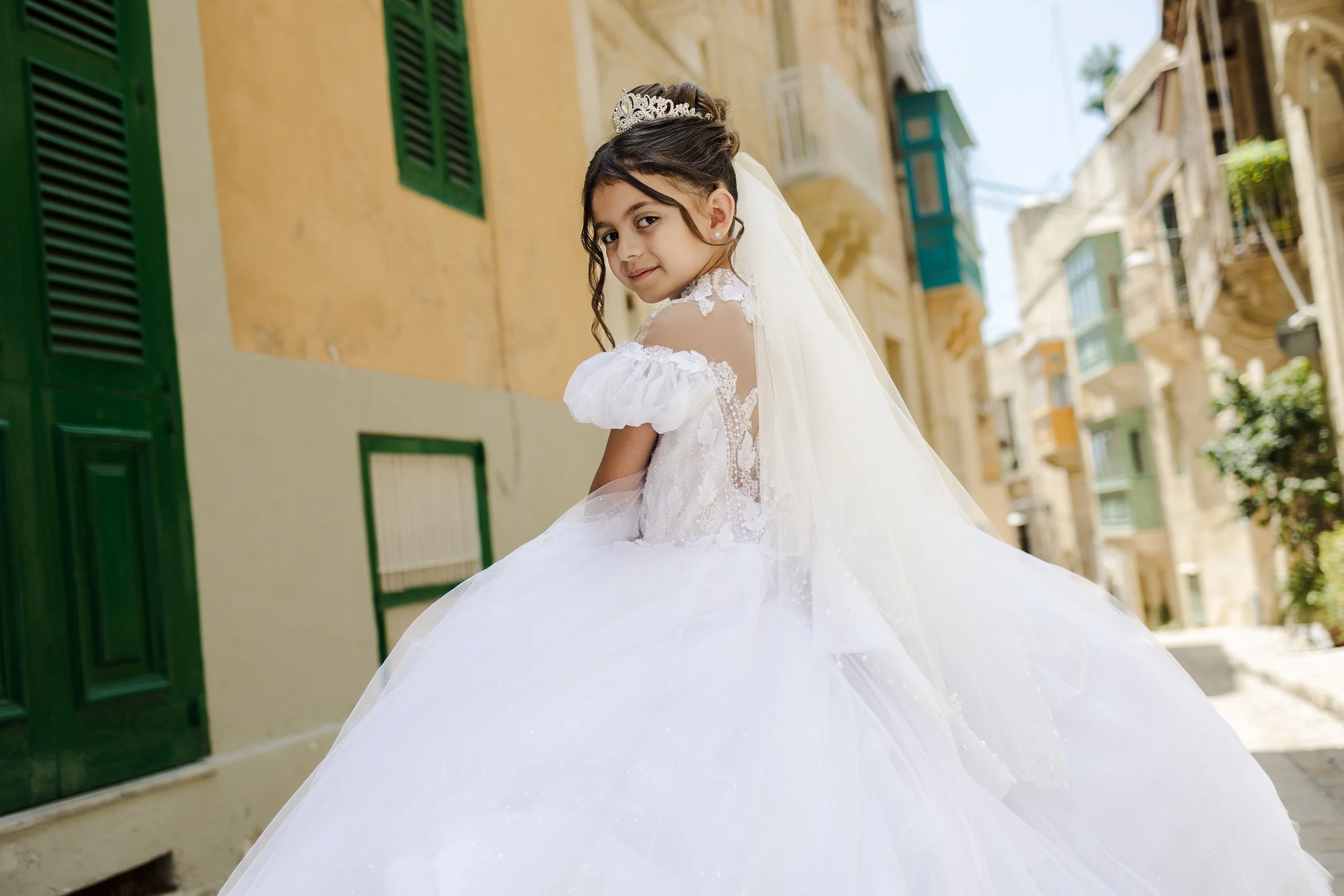 Young girl in a white wedding dress and veil, standing on a colorful urban street, smiling over her shoulder.