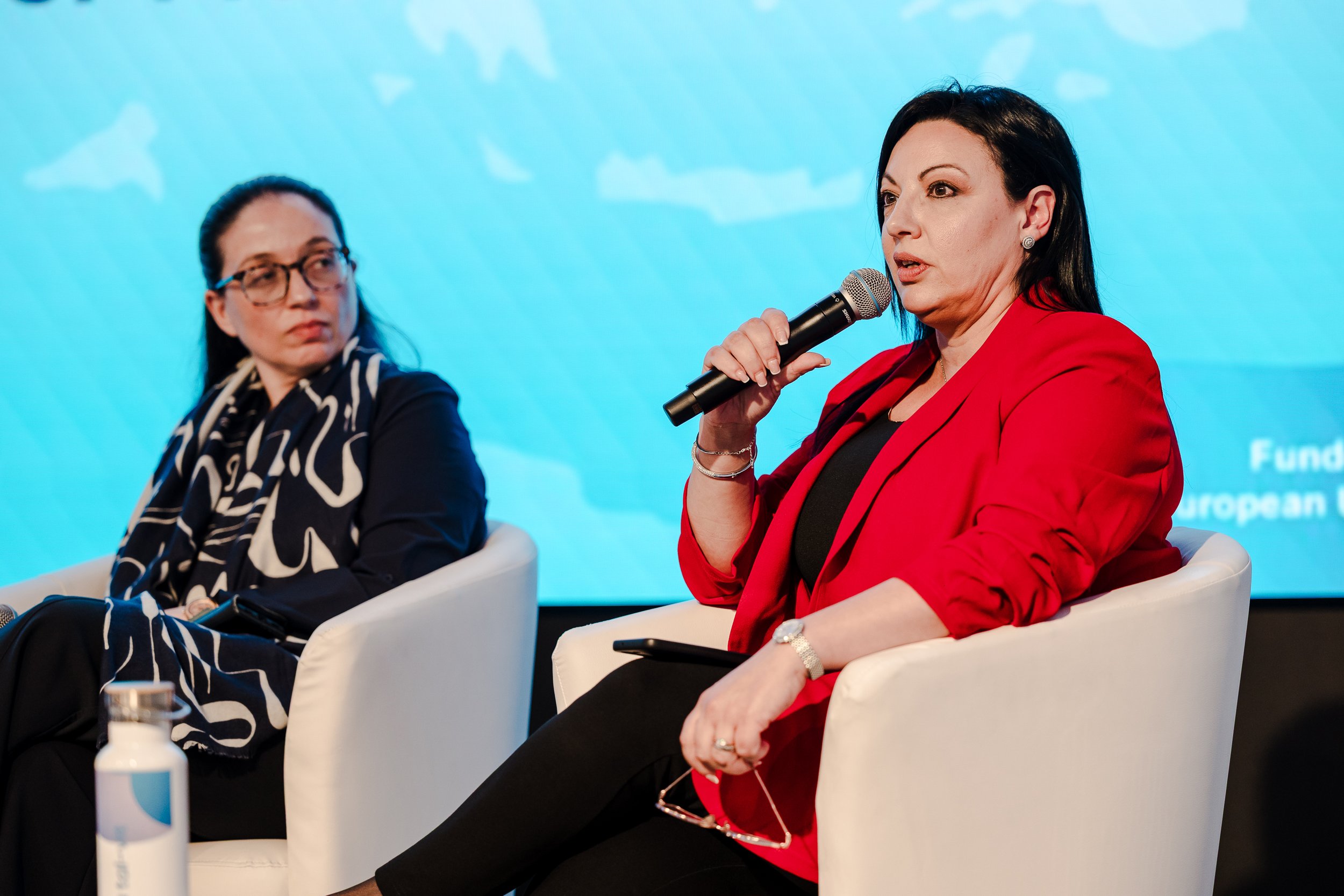 Two women seated on white chairs during a panel discussion. The woman on the right, wearing a red blazer and black shirt, is holding a microphone and speaking. The woman on the left, dressed in black with a patterned scarf, is listening. A bottle of 