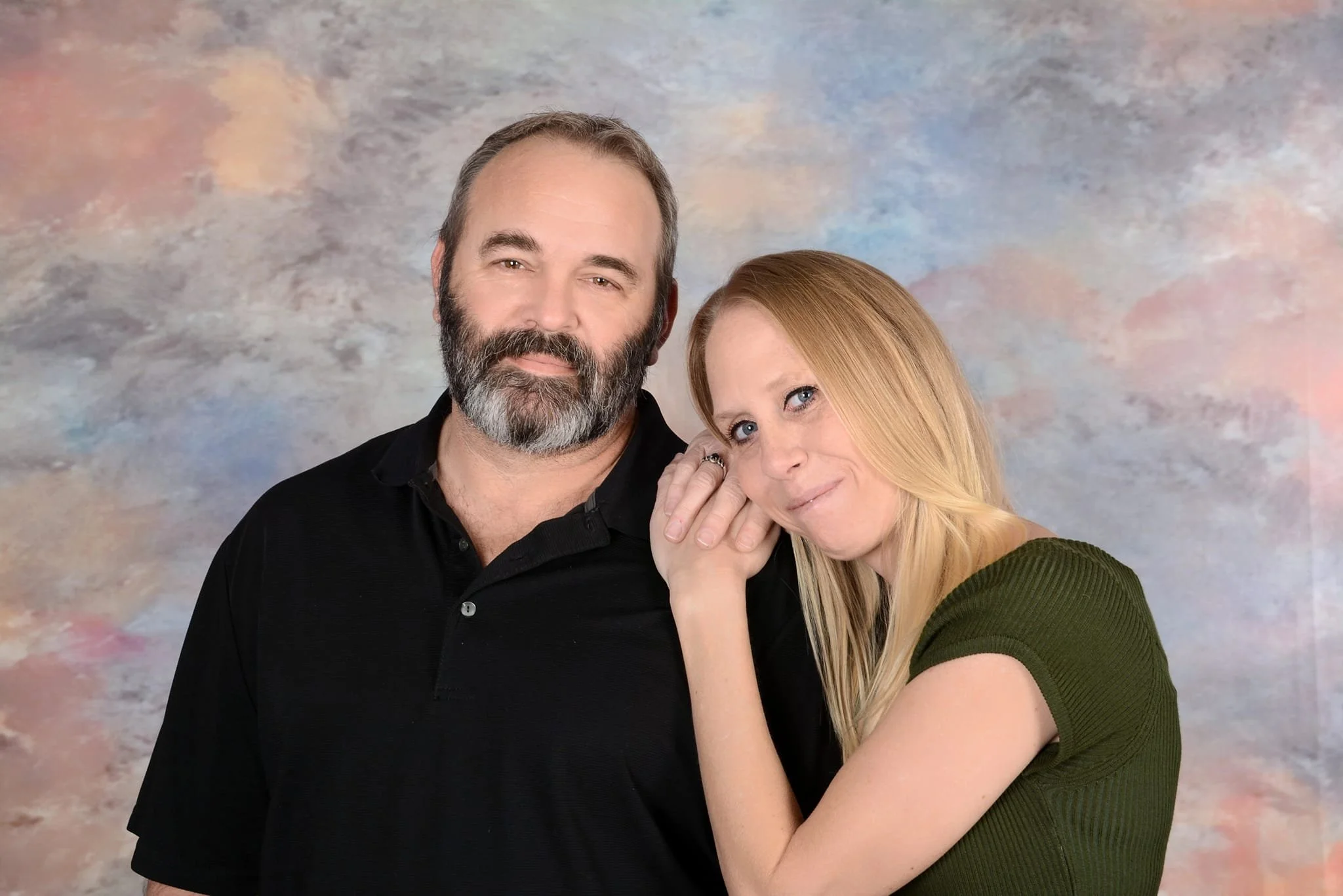 A middle-aged man with a beard and mustache, wearing a black shirt, standing next to a young woman with long blonde hair and blue eyes, dressed in a green top. They are posing closely together against a soft, multicolored background.
