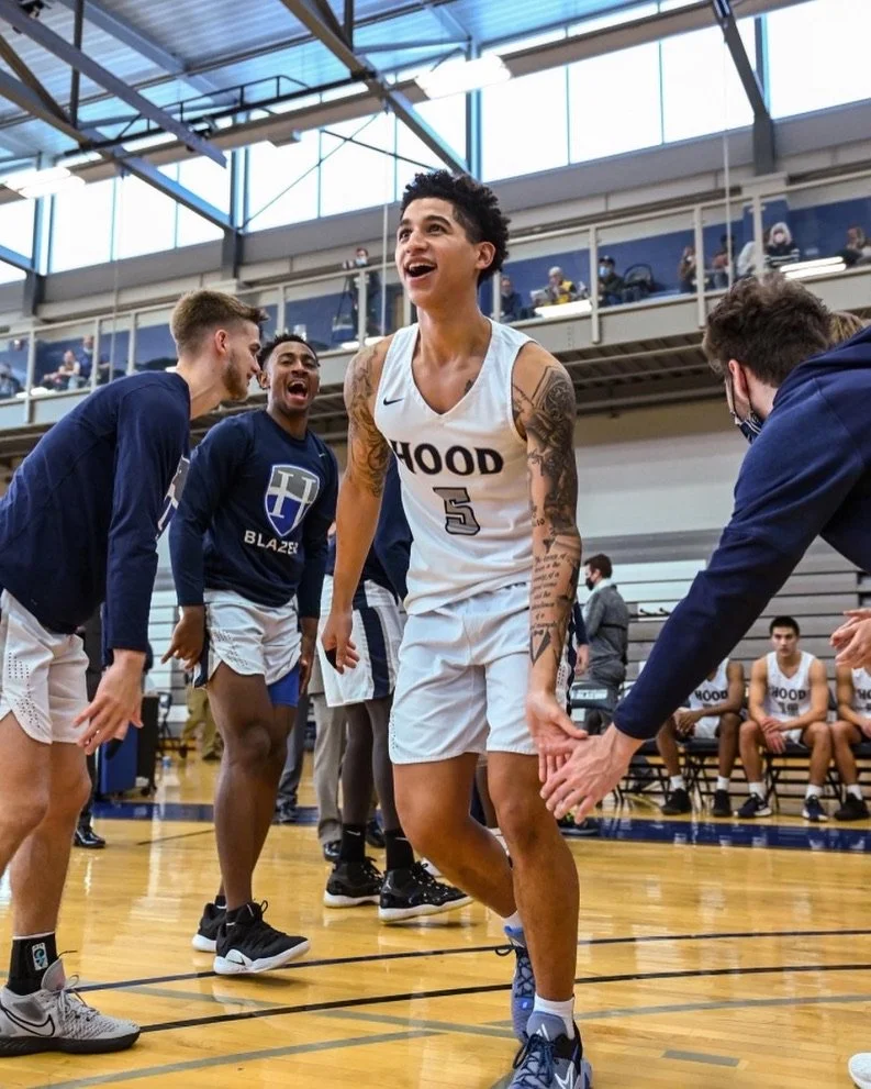 A basketball player wearing a white jersey with number 5 and the word 'HOOD' is smiling and high-fiving teammates on a court, with spectators watching from the stands.