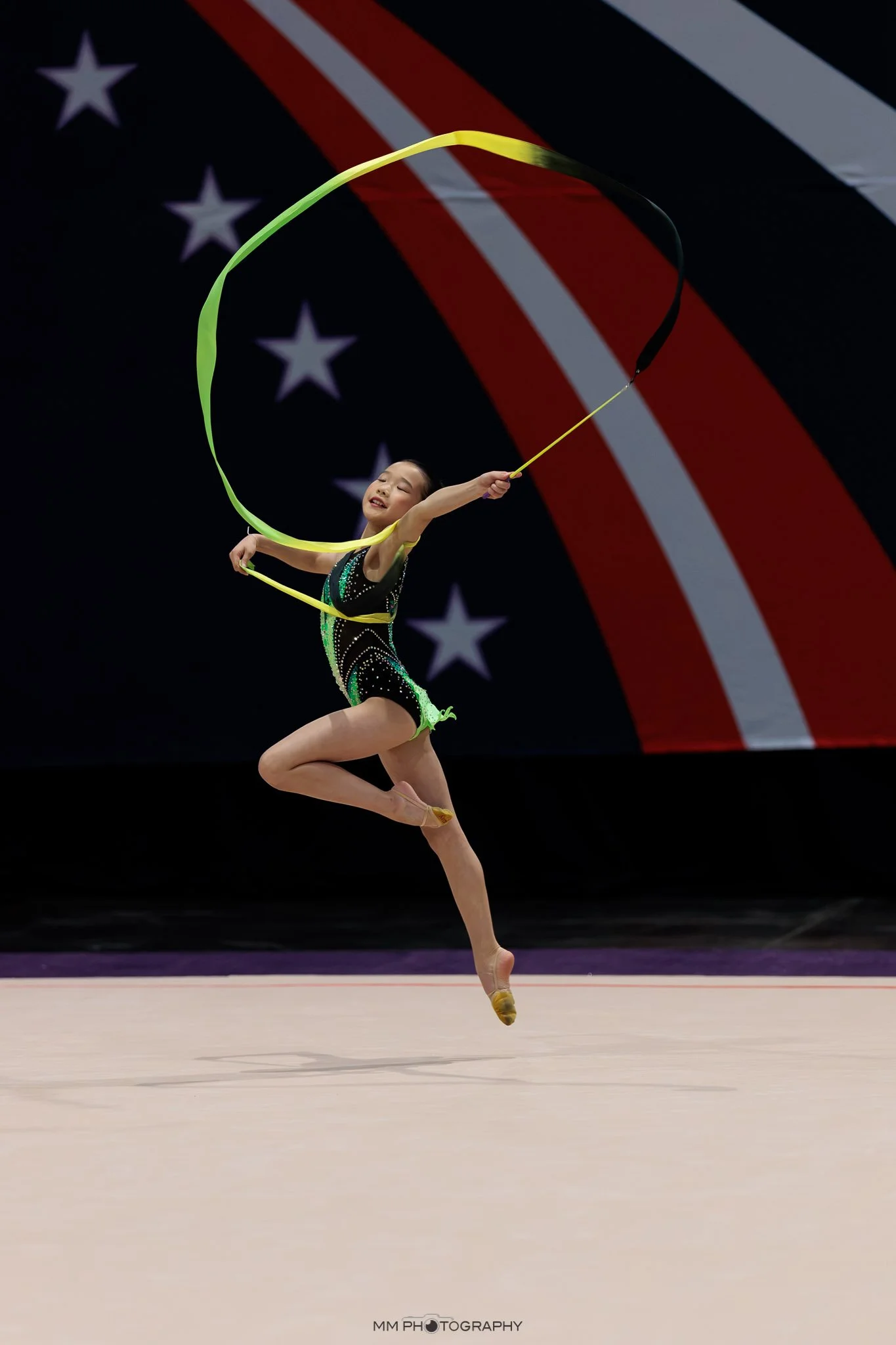 A young gymnast performing a jump during a gymnastics competition, with a large American flag in the background.