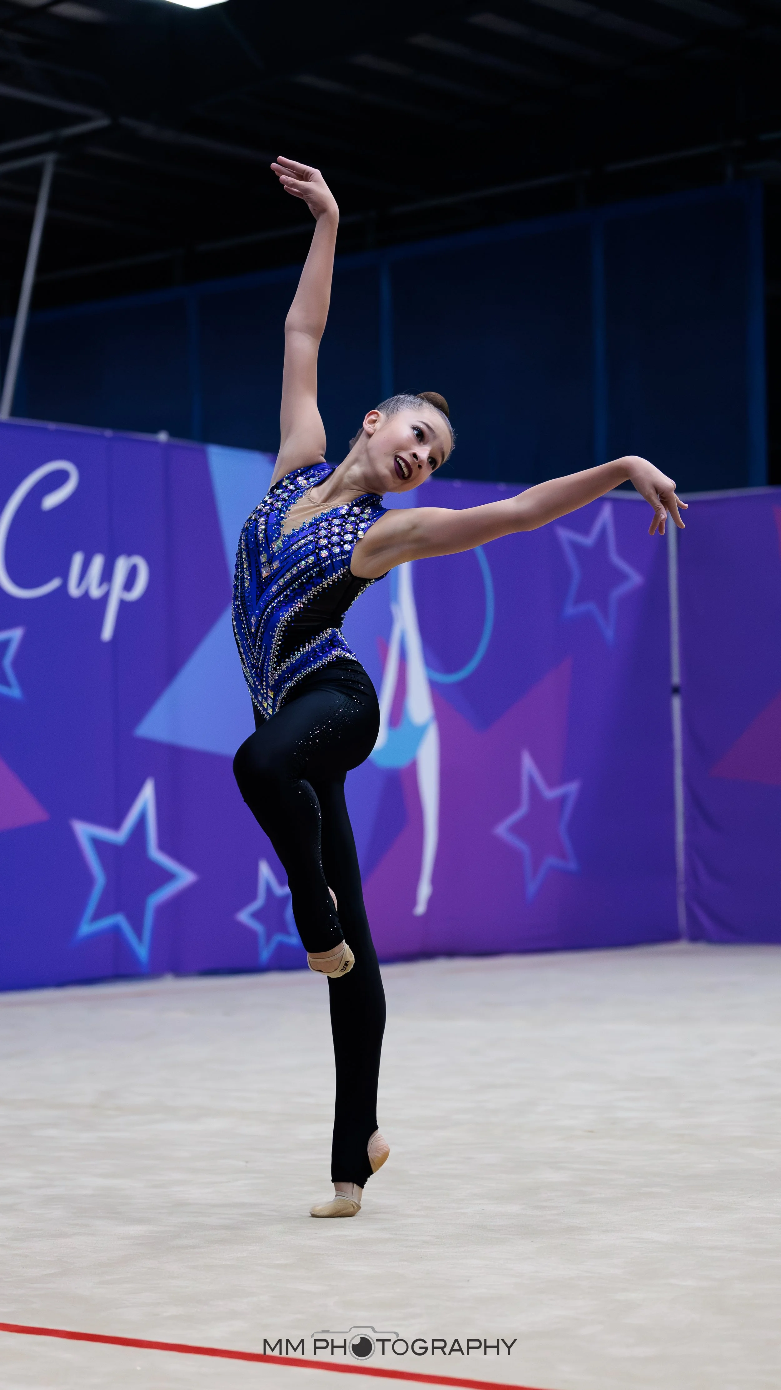 A female rhythmic gymnast in a sequined blue and black leotard performs a balance routine on the floor of a sports arena.