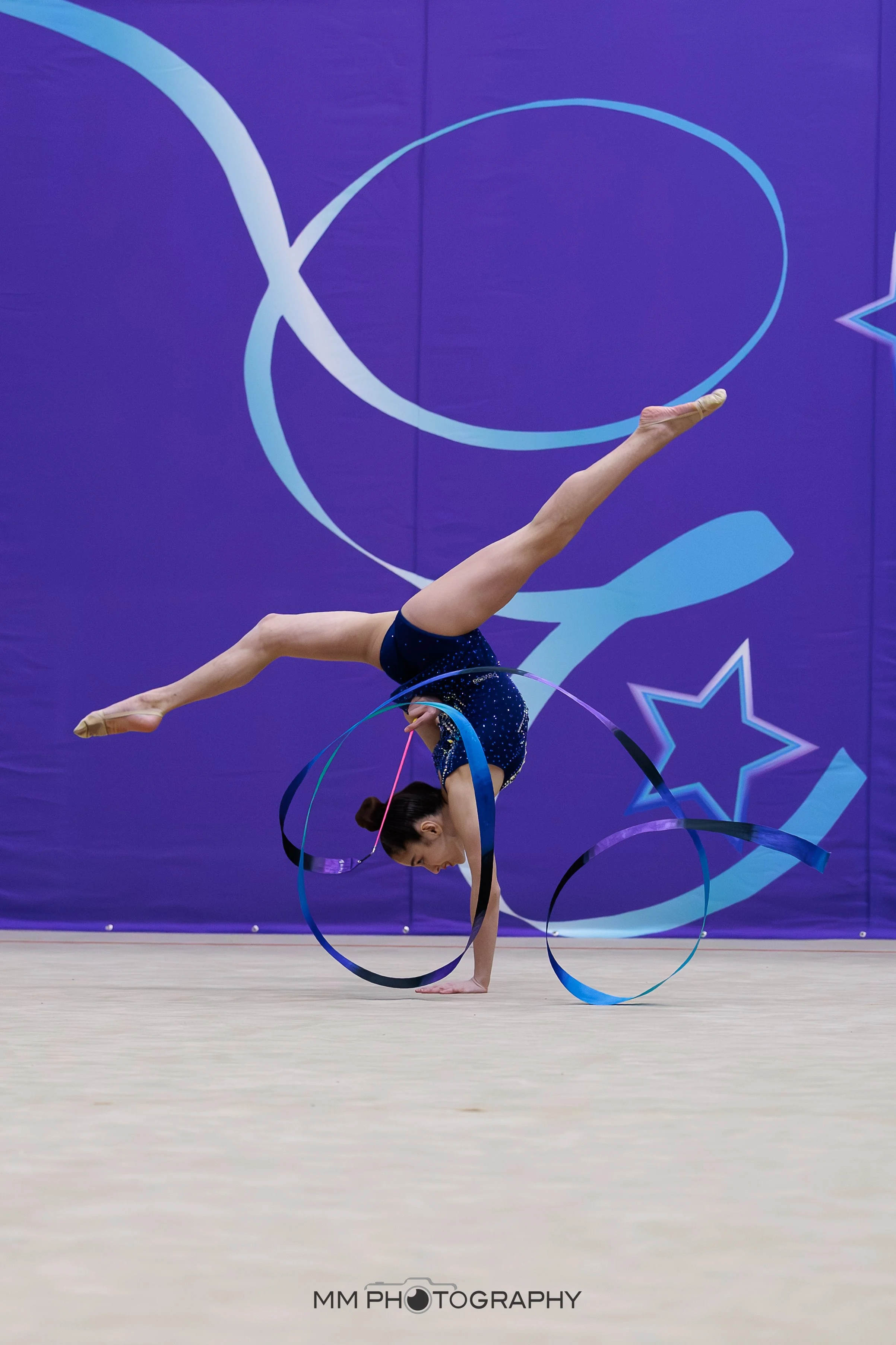 A gymnast performing a handstand routine with ribbons on a gymnastics floor in front of a purple backdrop with abstract designs.