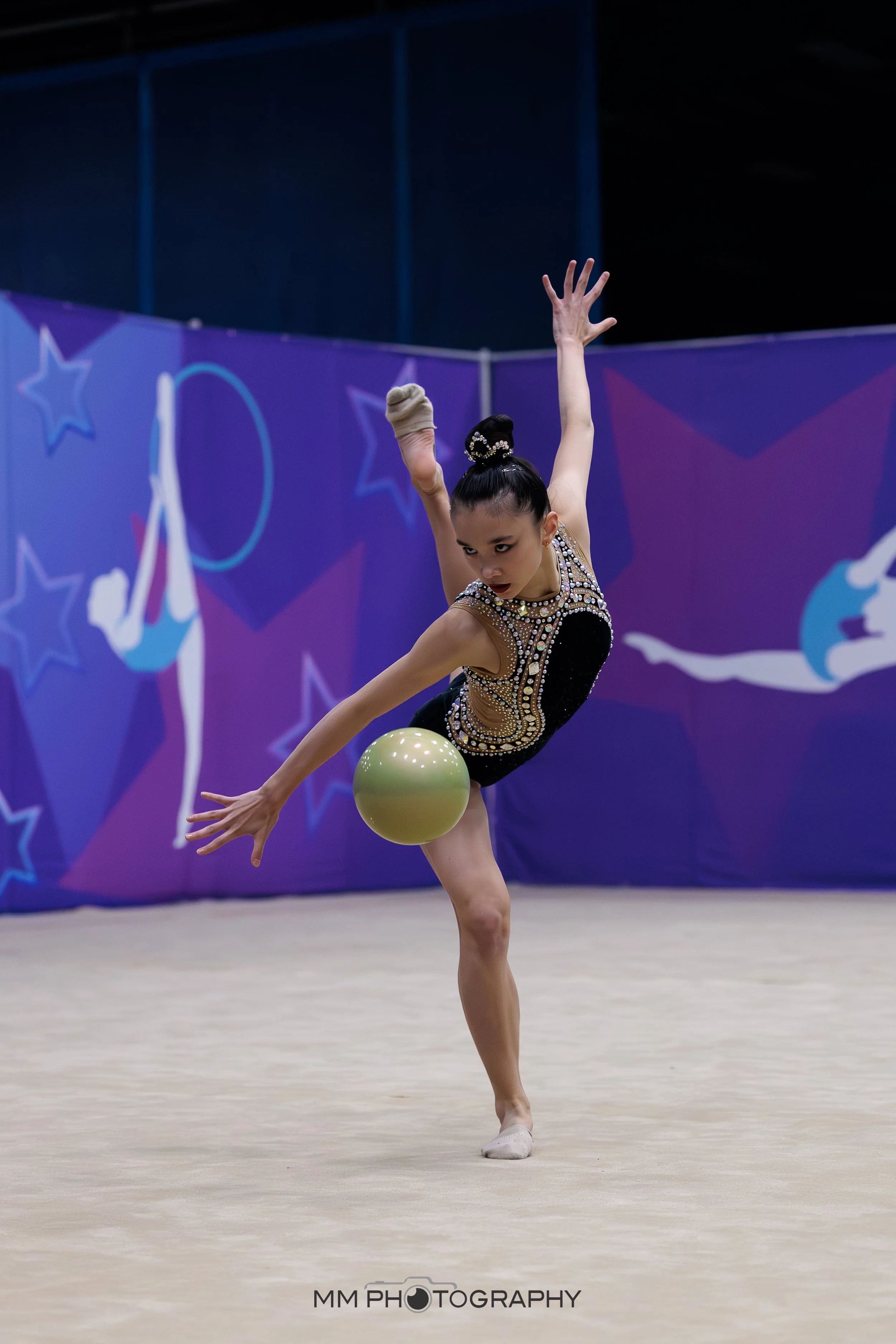 A rhythmic gymnast performing a move with a yellow ball during a competition, wearing a black and gold leotard, with a decorated hair bun, against a purple backdrop with star designs.