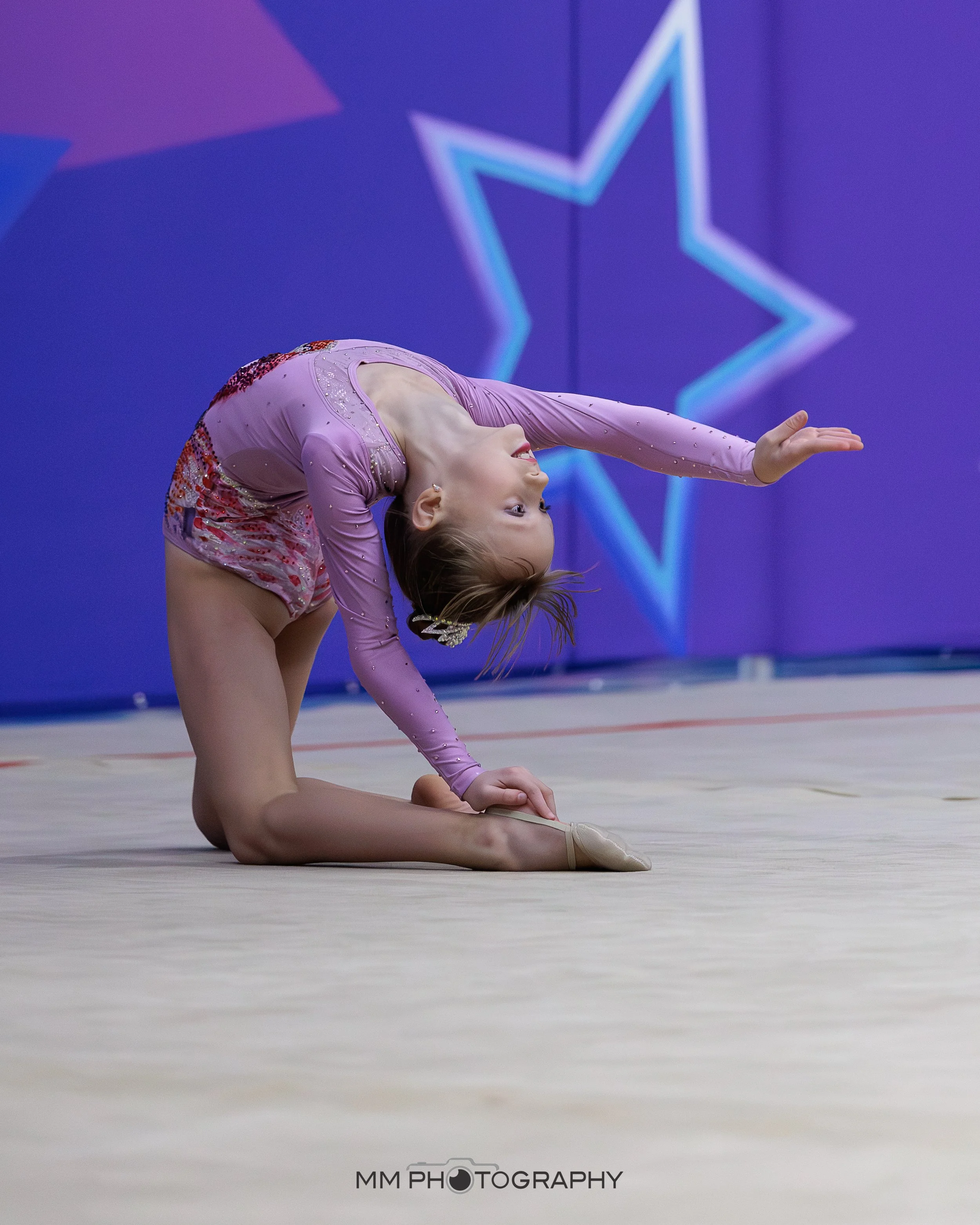 A young girl in a pink and purple figure skating dress performing a backbend on a beige skating rink with a purple and blue Background featuring a neon blue star outline.