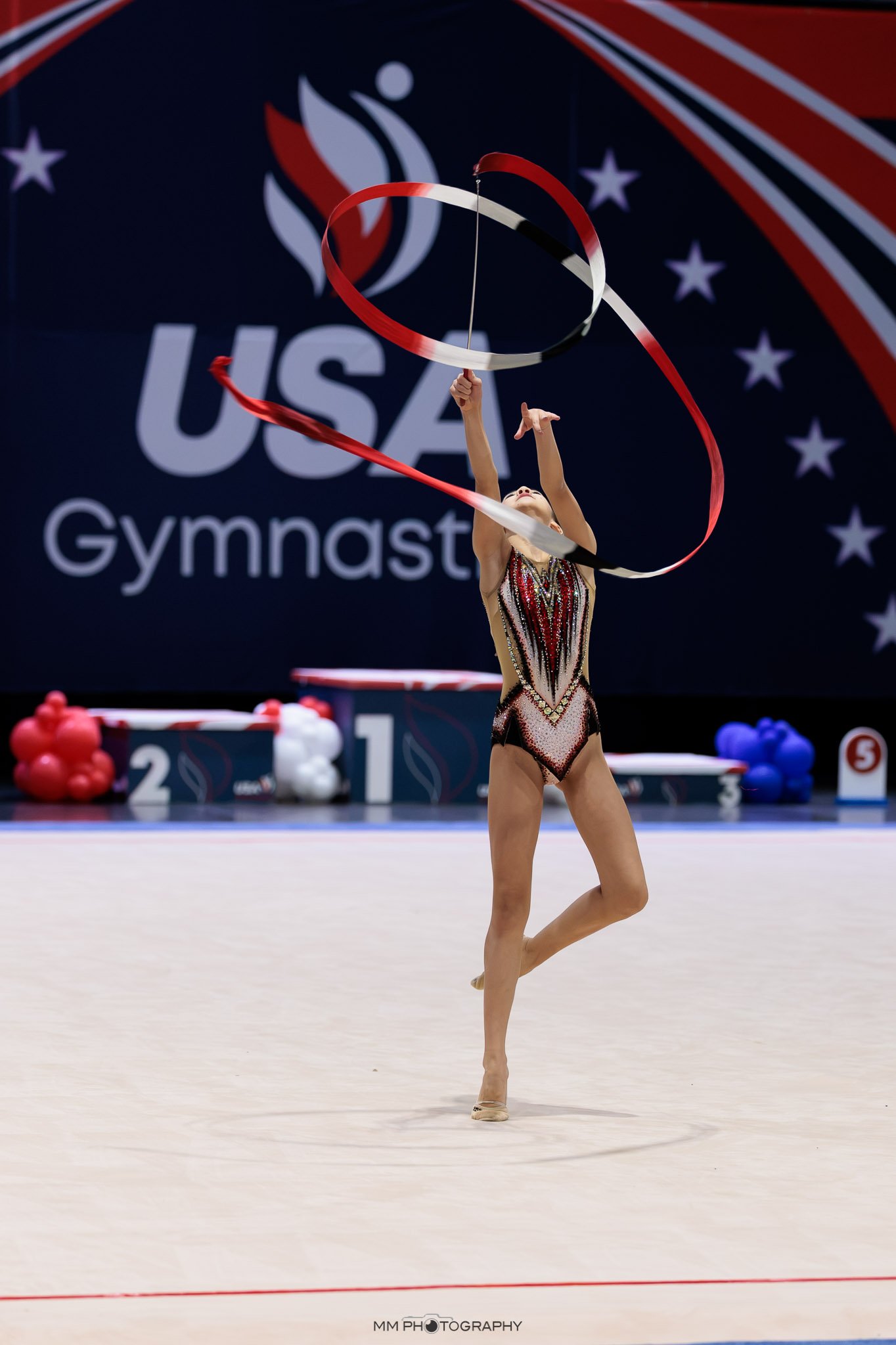 A gymnast performing a ribbon routine at a competition, with USA Gymnastics branding in the background.