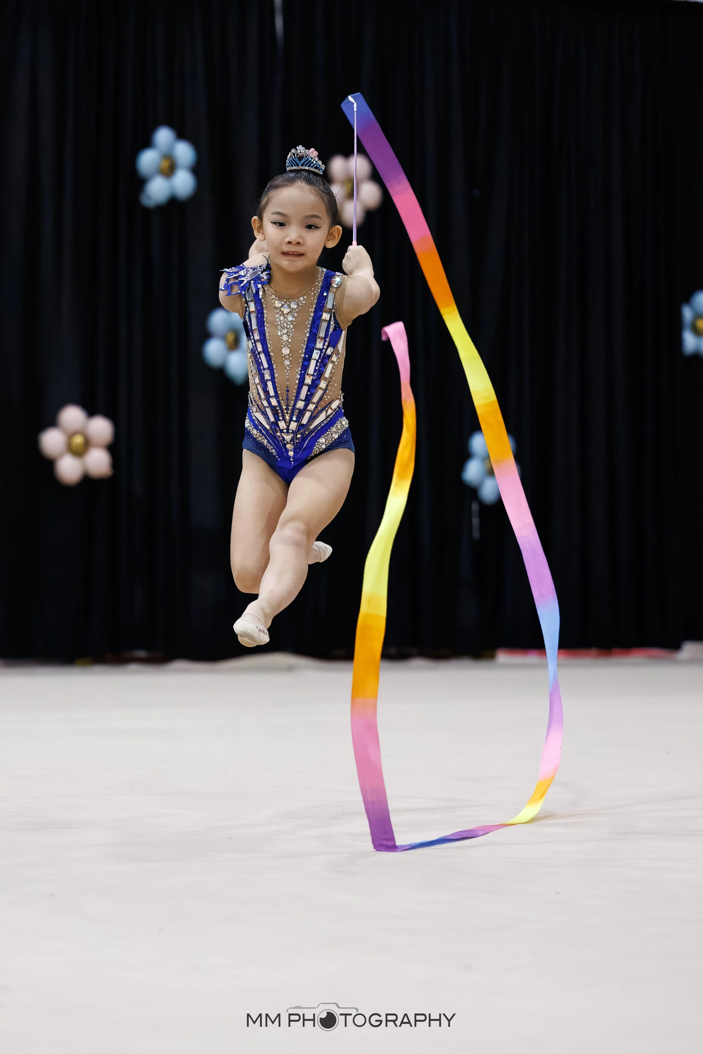 A young girl performing rhythmic gymnastics with a multicolored ribbon in an indoor gymnasium, wearing a blue and gold leotard with a tiara, in front of black curtains with decorative flowers.