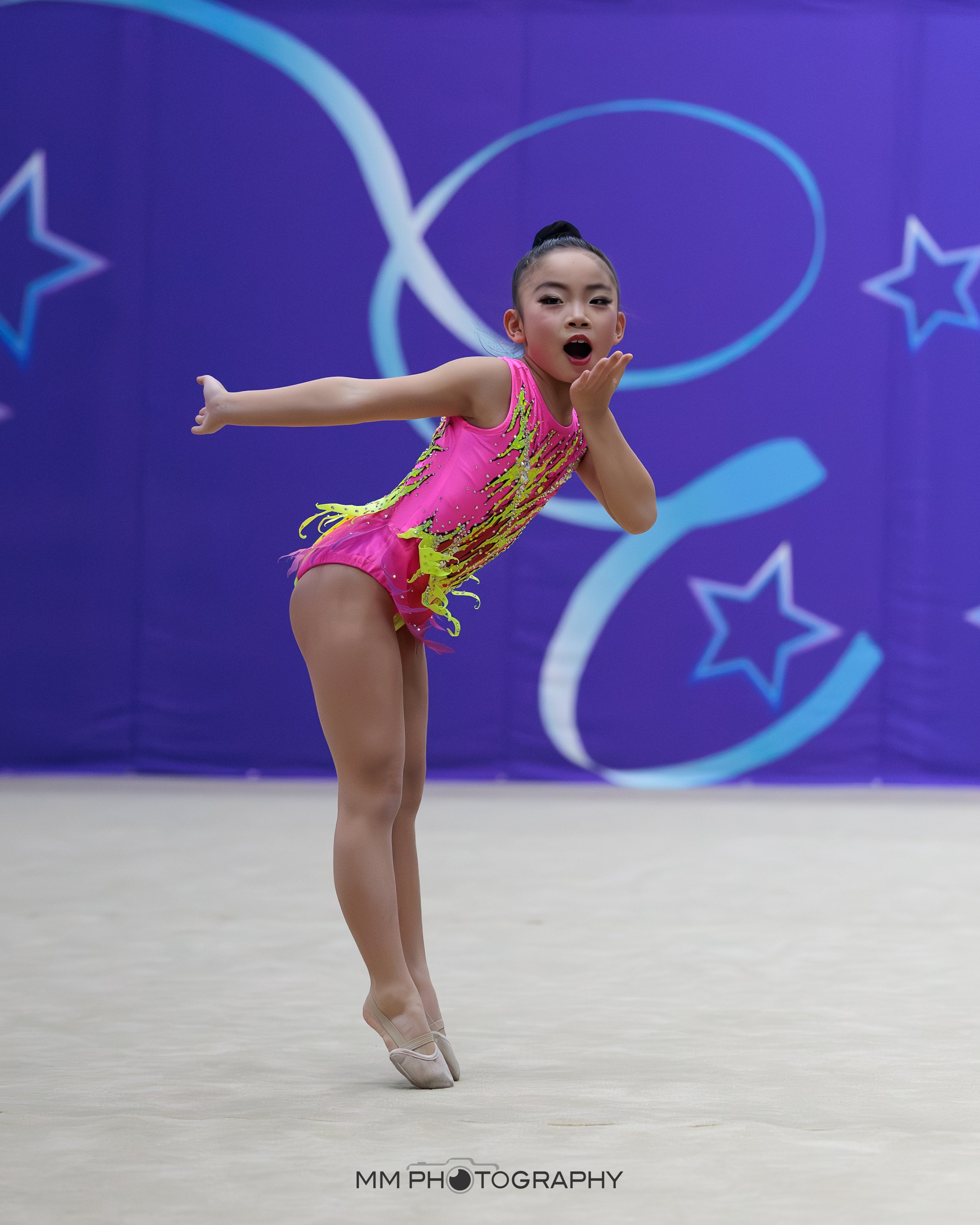 Young female gymnast performing a dance move on the floor during a competition, wearing a bright pink and yellow leotard, with a purple background decorated with star symbols.