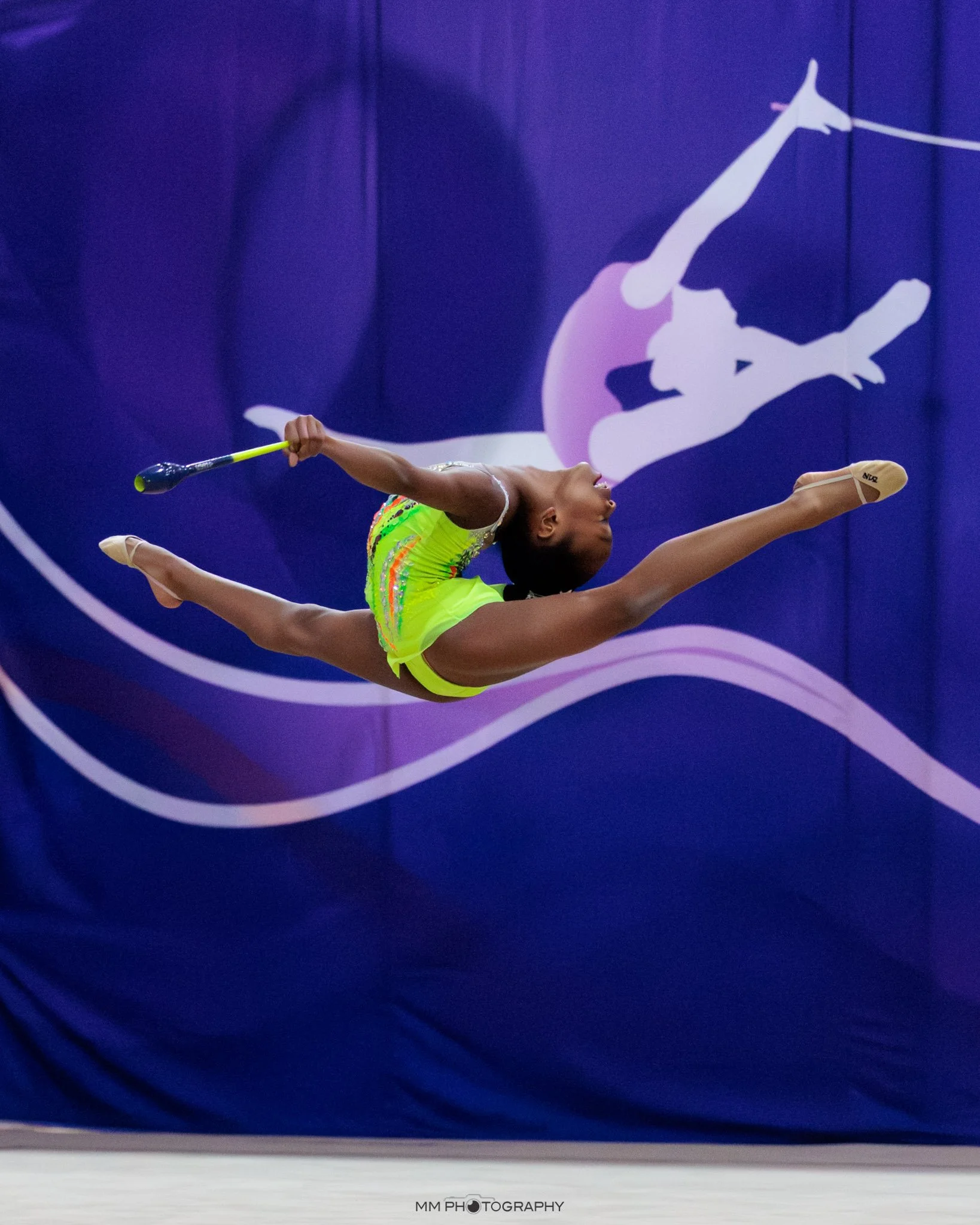 A young gymnast in a ballet pose, upside down, on a dark blue gymnastics floor with a purple background and a logo of a gymnast in silhouette.