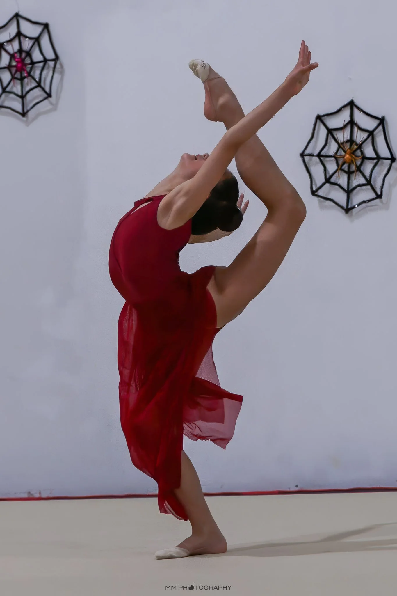 A rhythmic gymnast performing a pose on stage, wearing a red tutu and ballet slippers, with spider web decorations on the wall behind her.