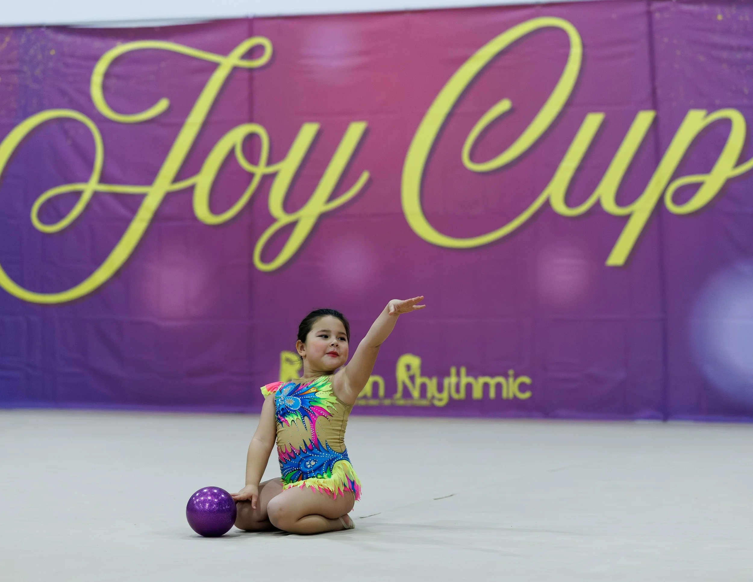 Young girl performing rhythmic gymnastics routine, sitting on the floor with one arm raised, dressed in a colorful leotard with vibrant patterns, purple ball beside her, in front of a purple banner with yellow cursive text.