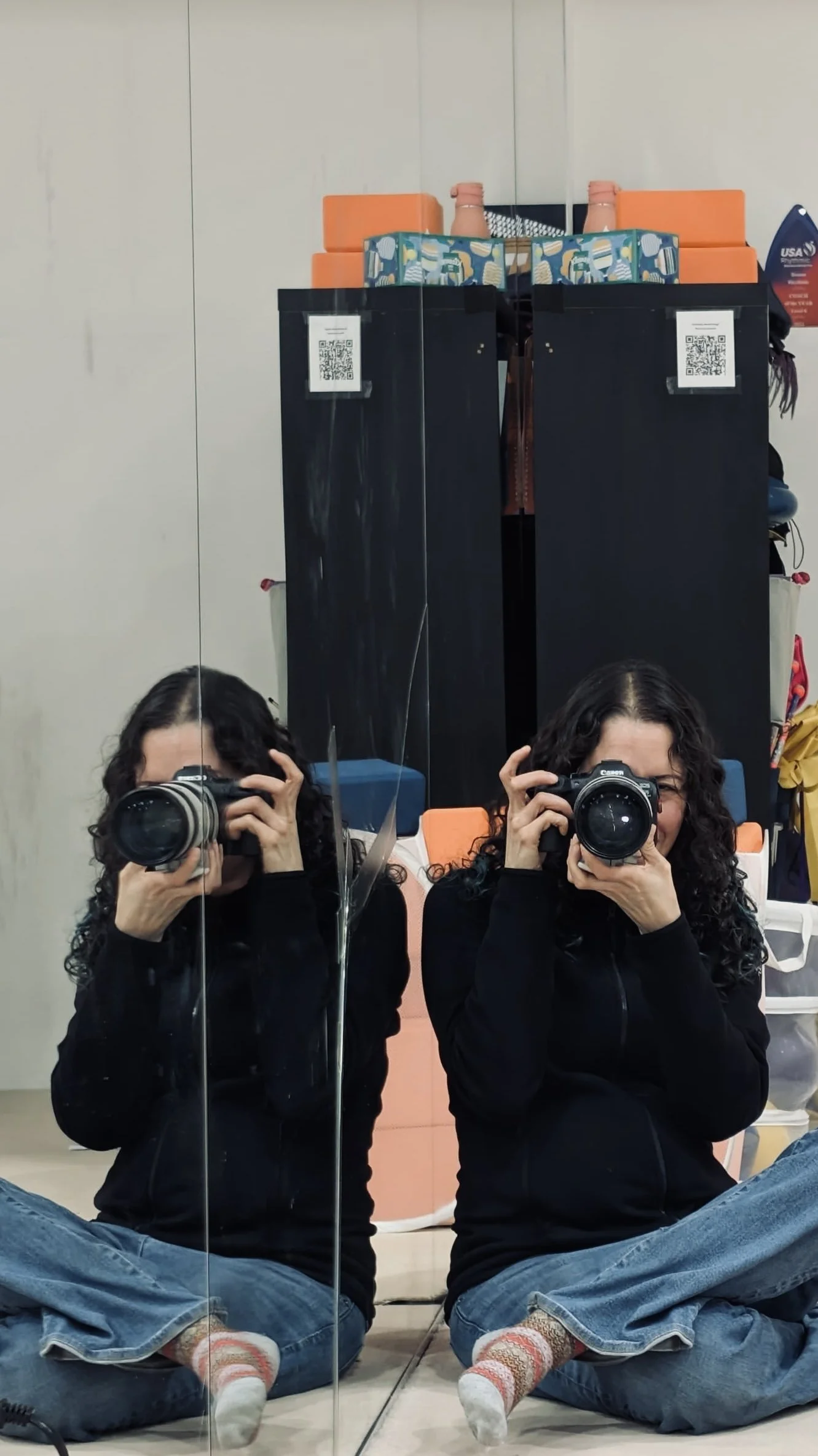 Two women with curly dark hair taking a mirror selfie with a camera, sitting cross-legged on the floor in front of a mirror, in a room with a black cabinet, orange cushions, and various objects on top.