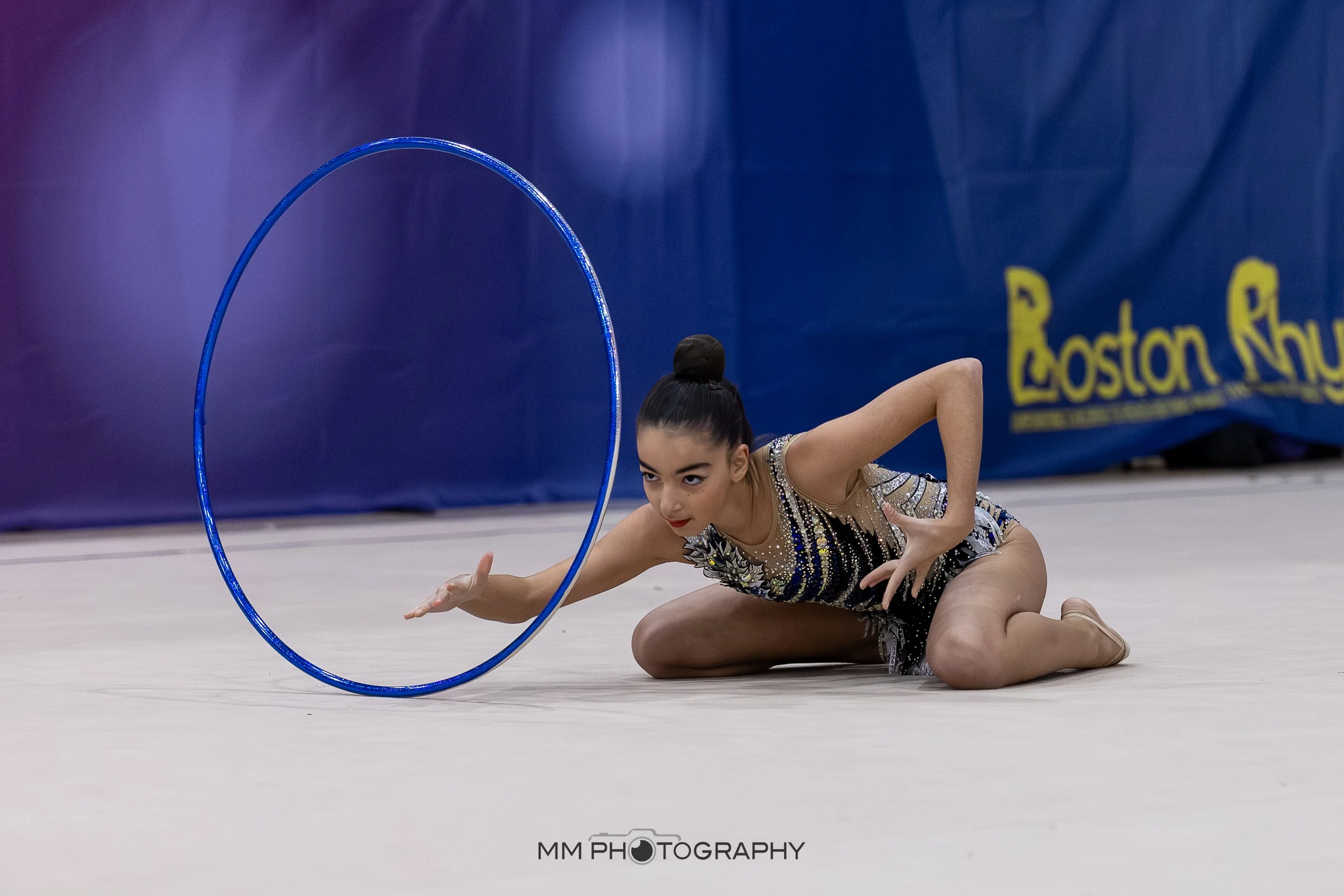 A young female gymnast performing a routine on the floor with a blue hoop in an indoor gymnasium. She is wearing a sparkly leotard and has her hair tied in a bun. There is a blue banner with yellow letters that say "Boston" in the background.