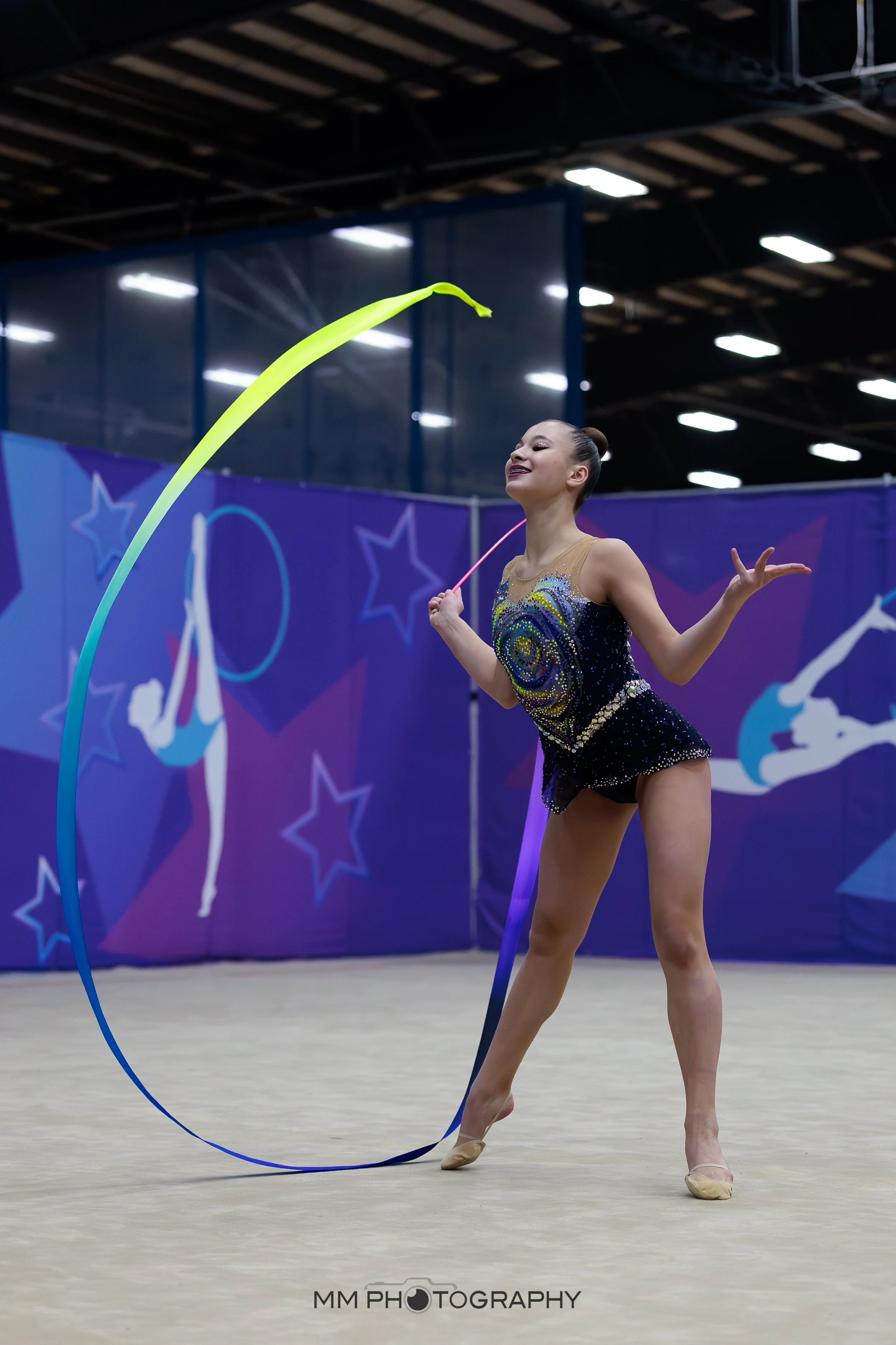 A rhythmic gymnast performing with a yellow ribbon in an indoor arena, wearing a sequined leotard and beige shoes.