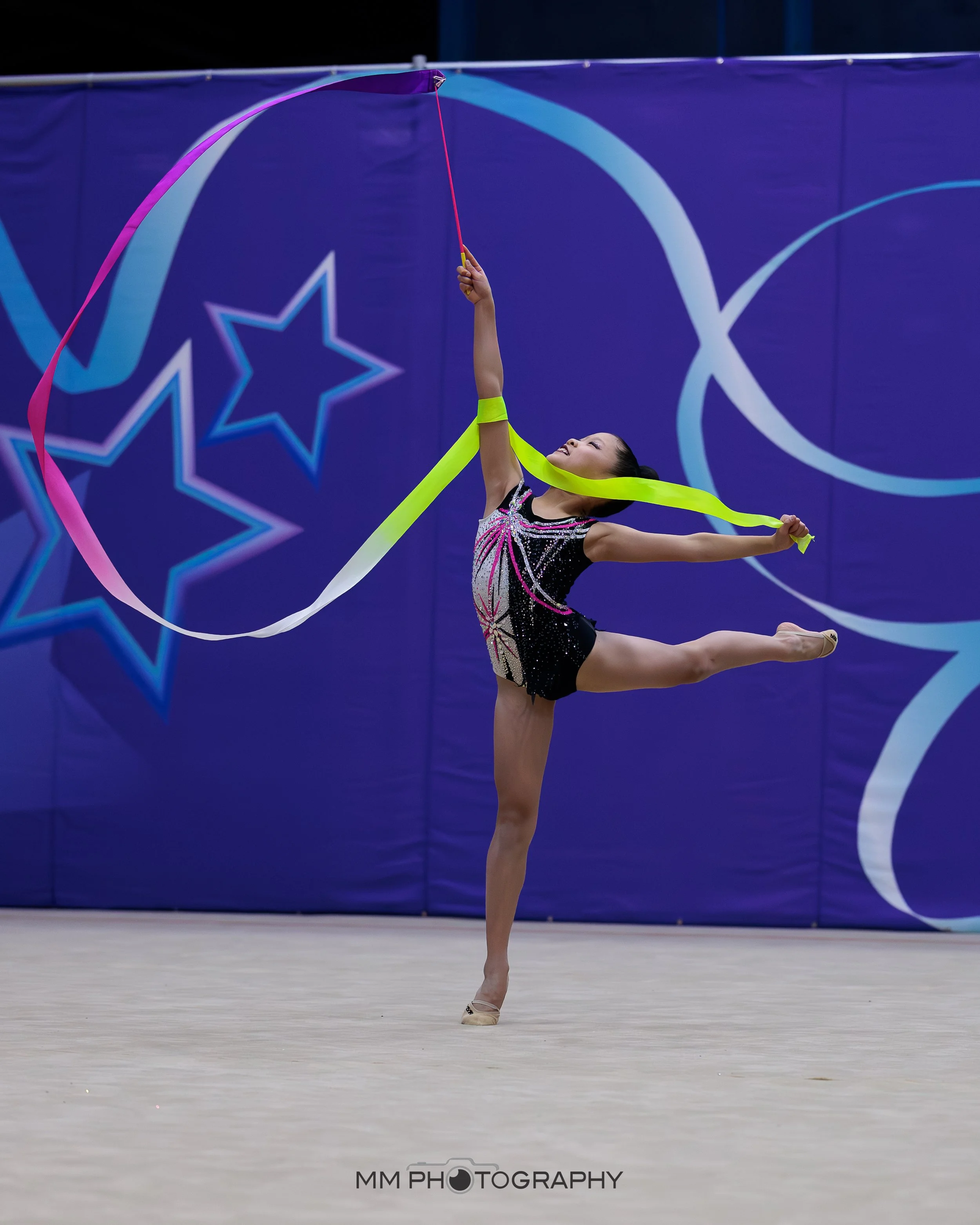 A female rhythmic gymnast performing a pose with a pink and yellow ribbon in front of a blue backdrop with star designs, wearing a sparkly leotard.