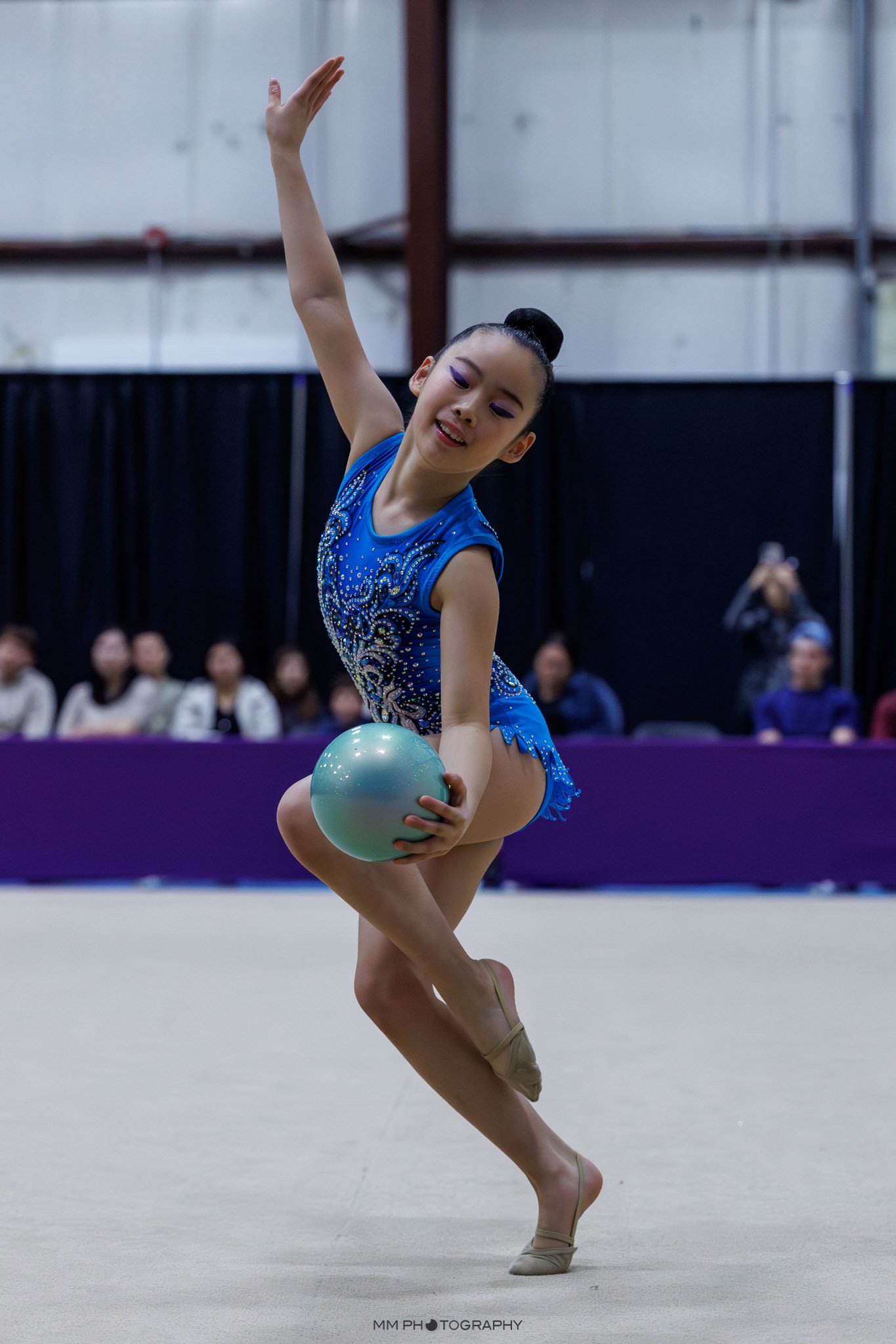 A young female gymnast in a blue and silver leotard is balancing on her right foot, holding a light blue ball, with her left leg raised and bent, and her arms extended elegantly in a gymnastics routine. She has black hair styled in a high bun and is 