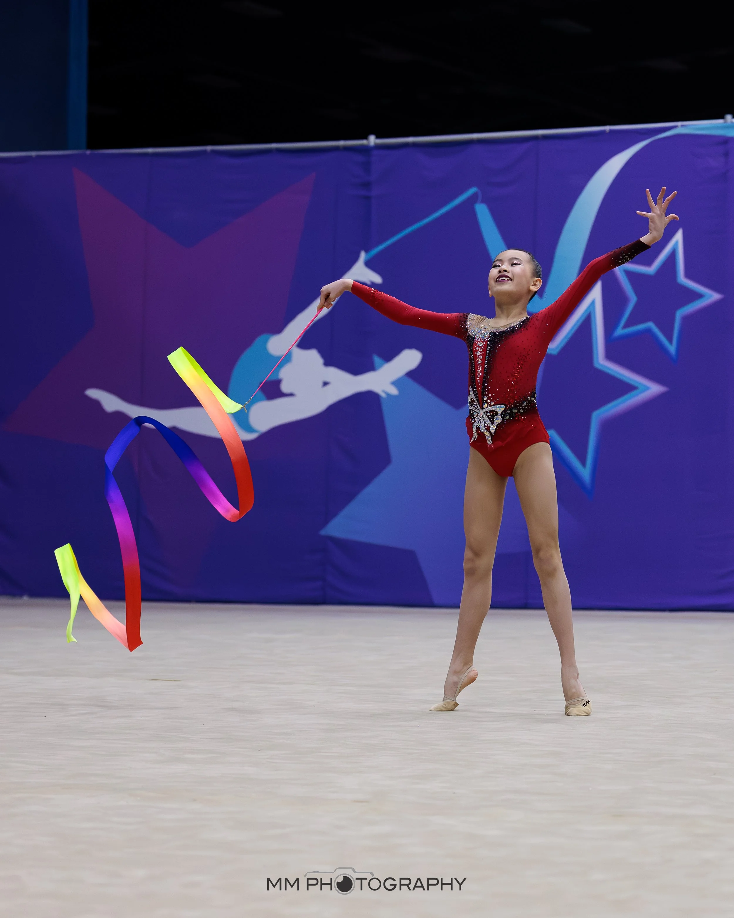 A young female gymnast in a red and black leotard performing with colorful ribbons during a gymnastics competition.