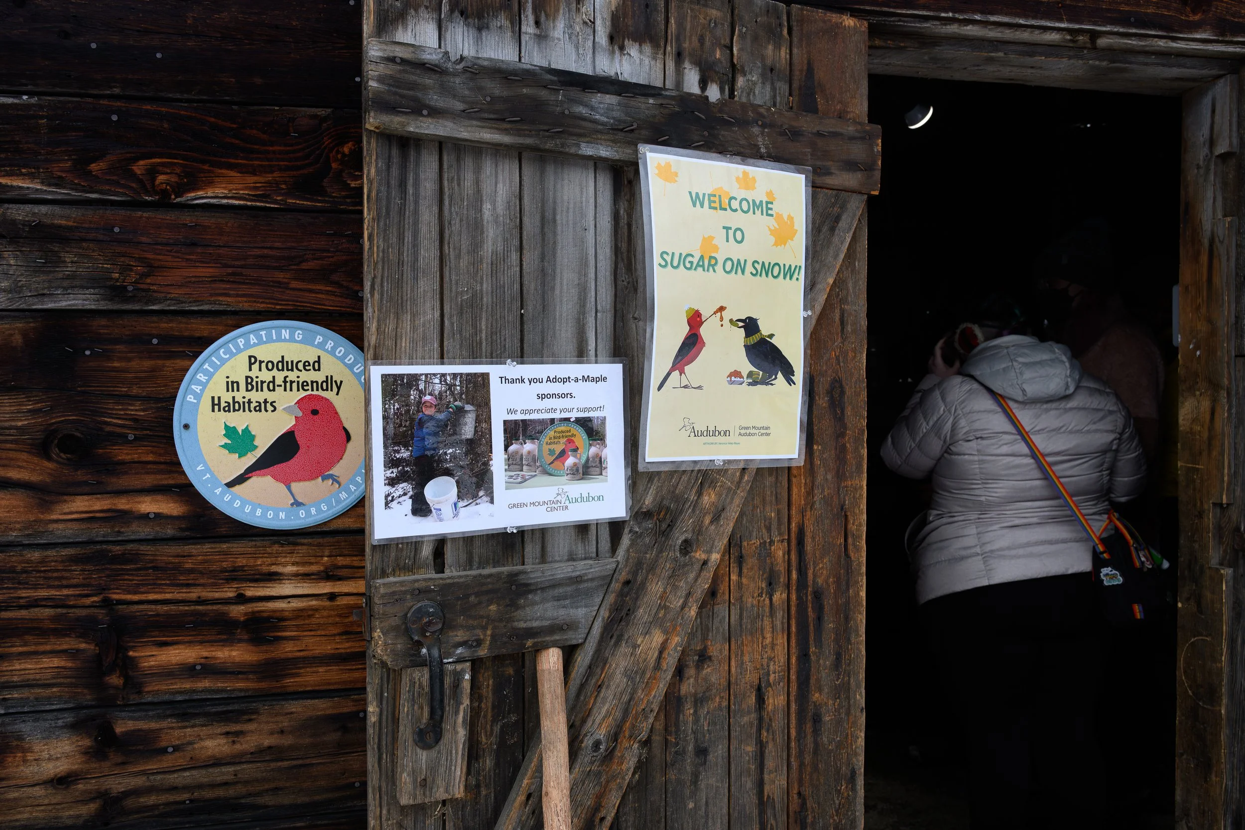 People entering a wooden barn with signs about bird habitats and a welcome message for snow sugar event.