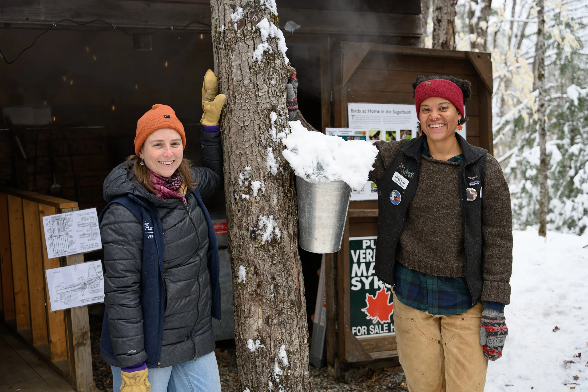 Two women standing outdoors in winter, smiling, with one woman touching a snow-covered tree and the other standing nearby. They are dressed warmly, wearing coats, hats, and gloves.