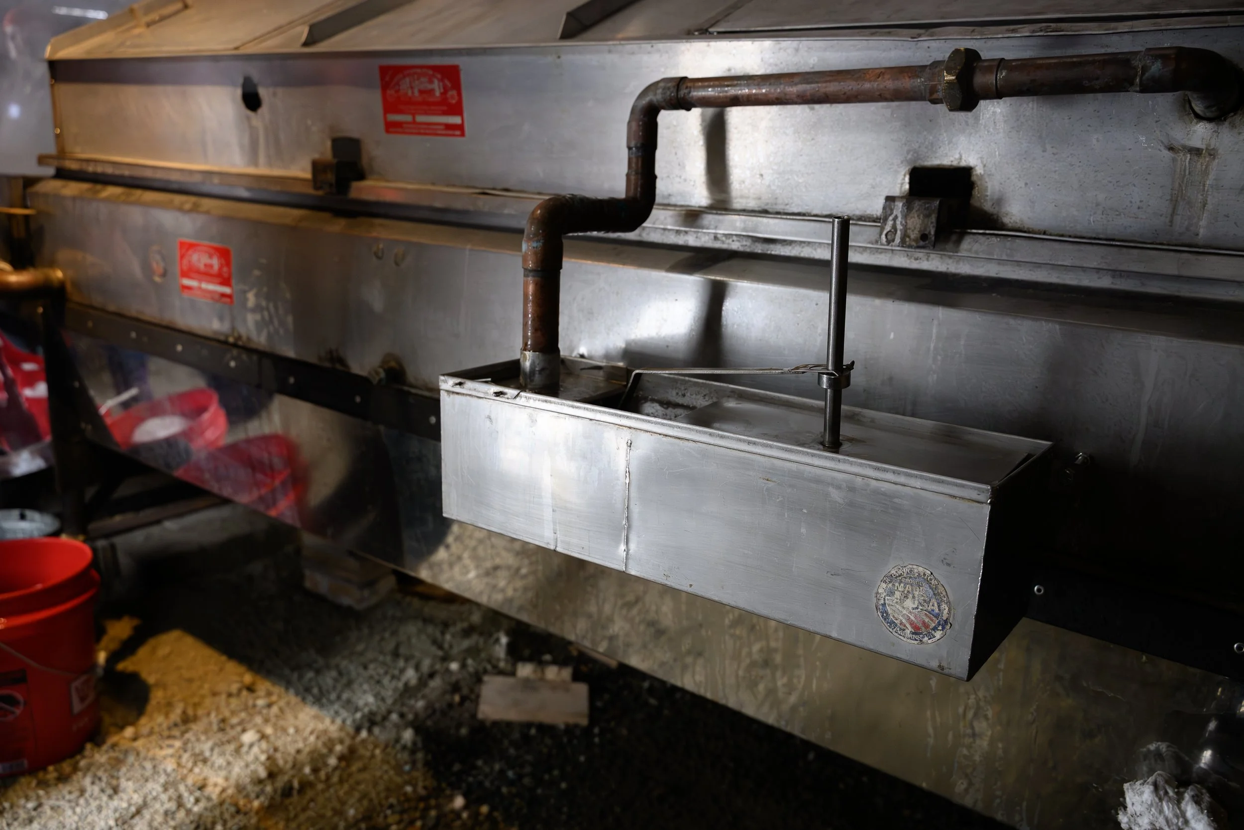 A metal food warming or cooking unit with pipes and a faucet, located outdoors on dirt ground with some red buckets nearby.