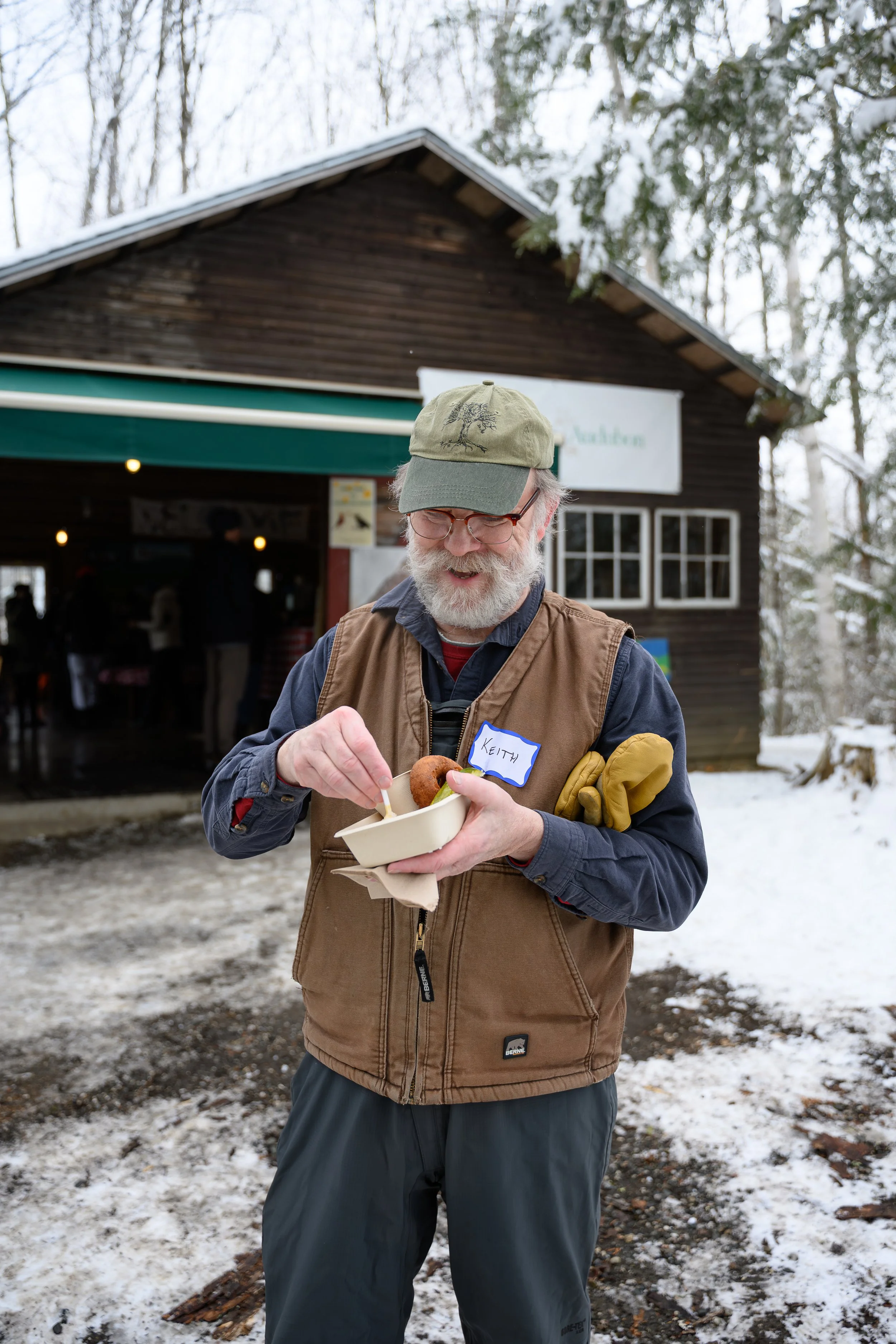 An older man with a white beard, glasses, wearing a tan vest over a blue shirt, and a green cap, standing outside in a snowy environment. He is holding a paper food container and a fork, smiling. There is a wooden building with a green awning and a s
