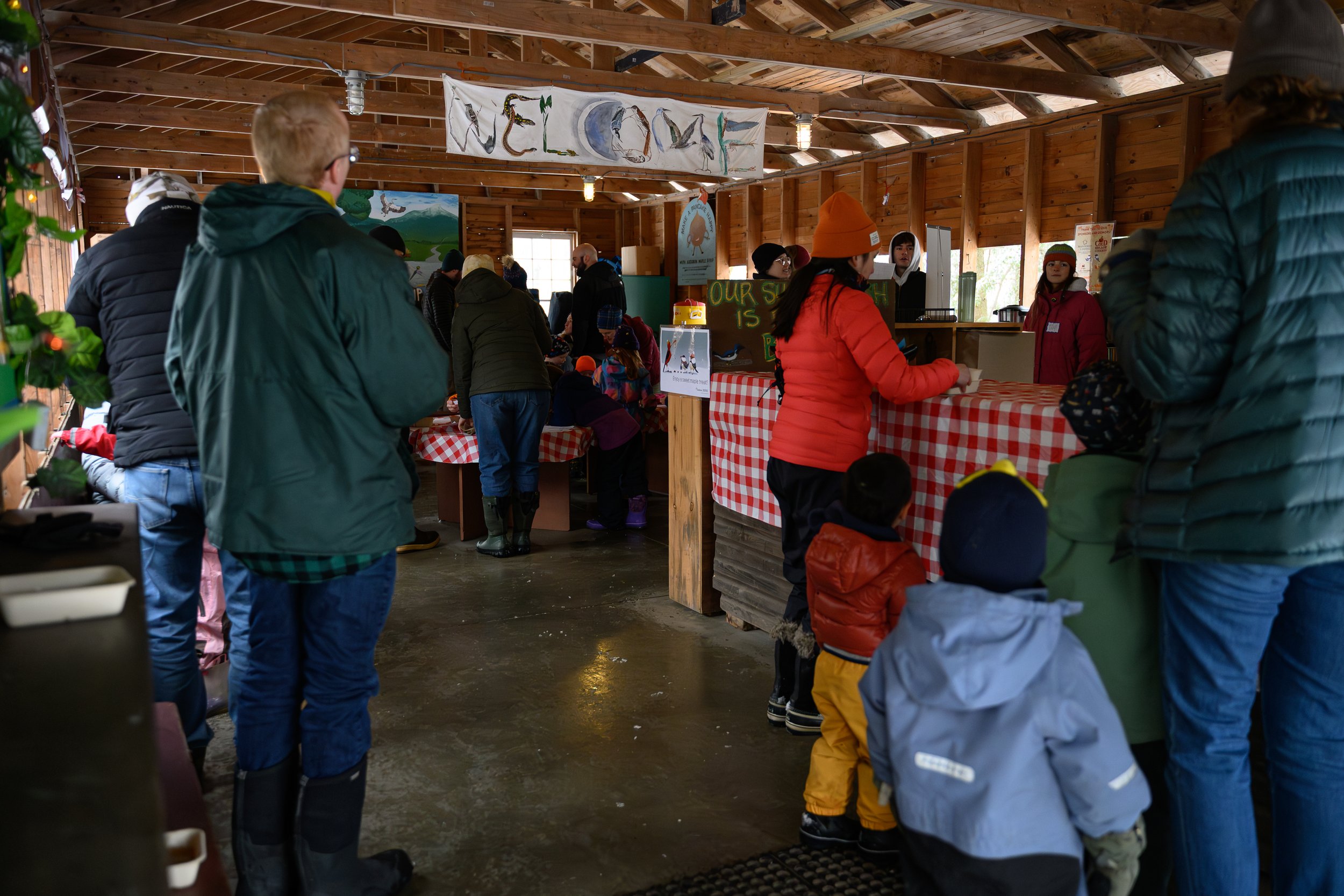 People inside a wooden building with a checkered tablecloth, some waiting in line, and a banner hanging from the ceiling, possibly at an event or market.