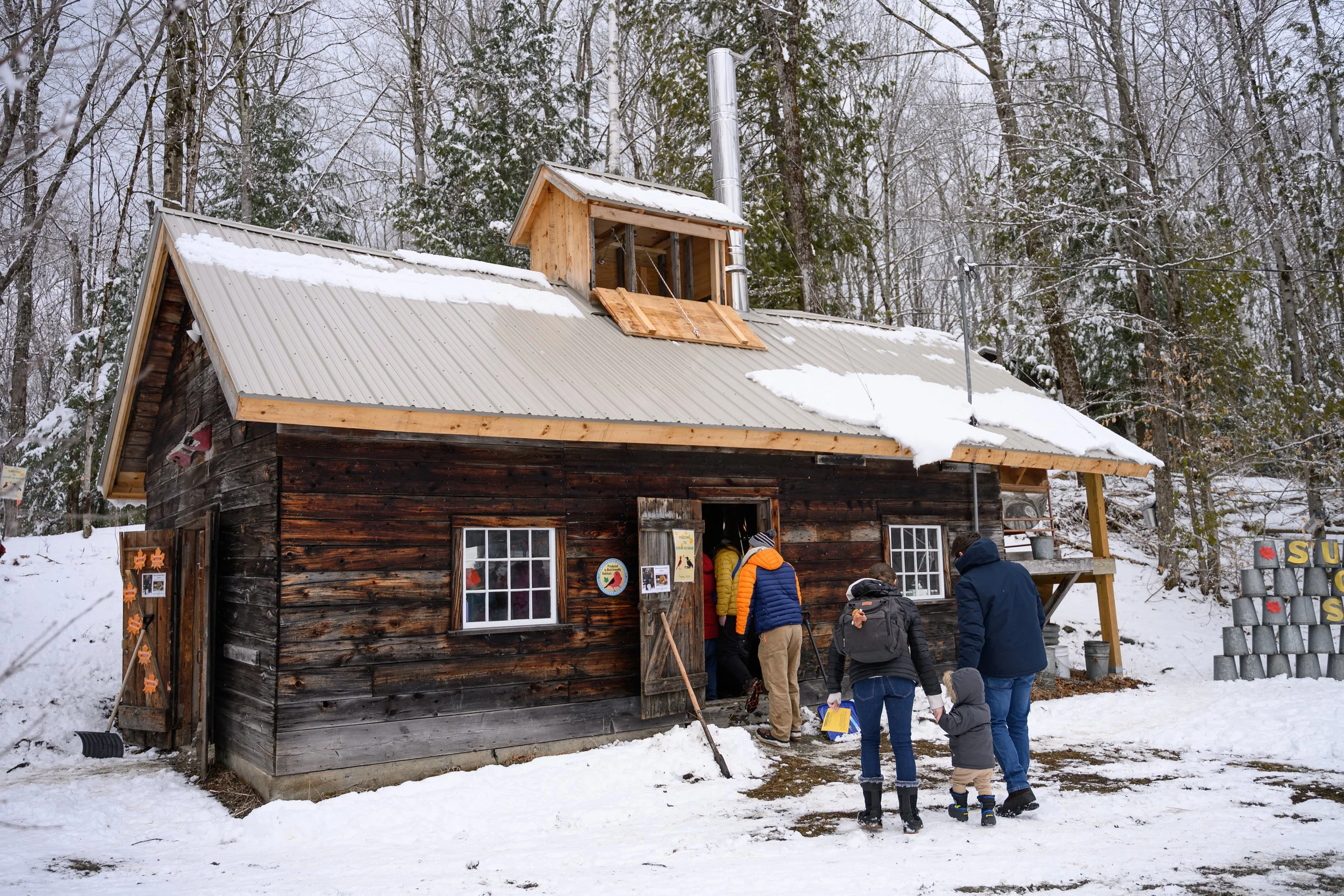 A group of people in winter clothing standing outside a rustic wooden building with a snow-covered sloped metal roof and a small dormer window under construction, surrounded by snow and bare trees.