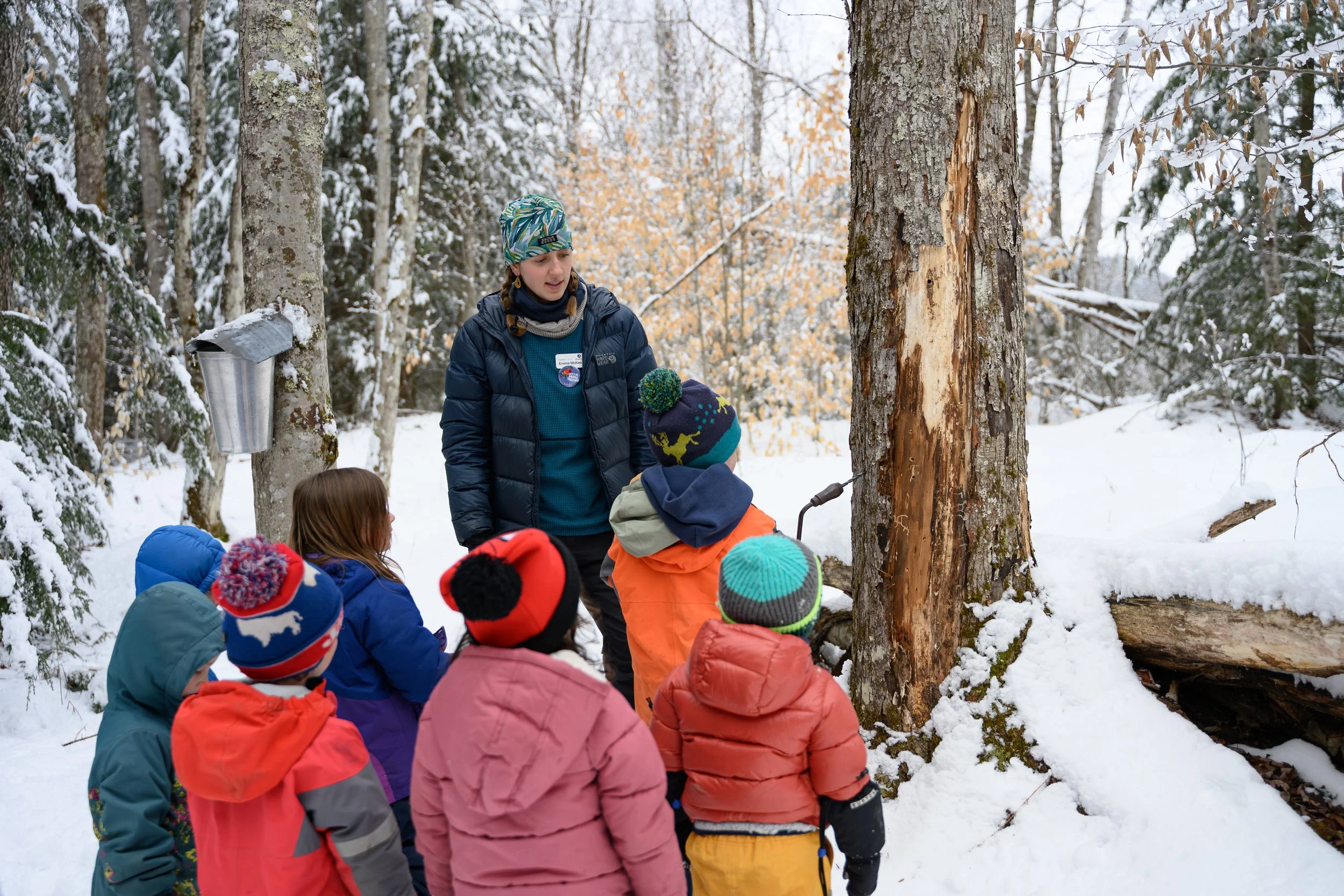 A woman leading a group of children in an outdoor educational activity in a snow-covered forest during winter. The woman is wearing a blue jacket and a colorful headband, and the children are dressed in colorful winter coats and hats. The woman is ex
