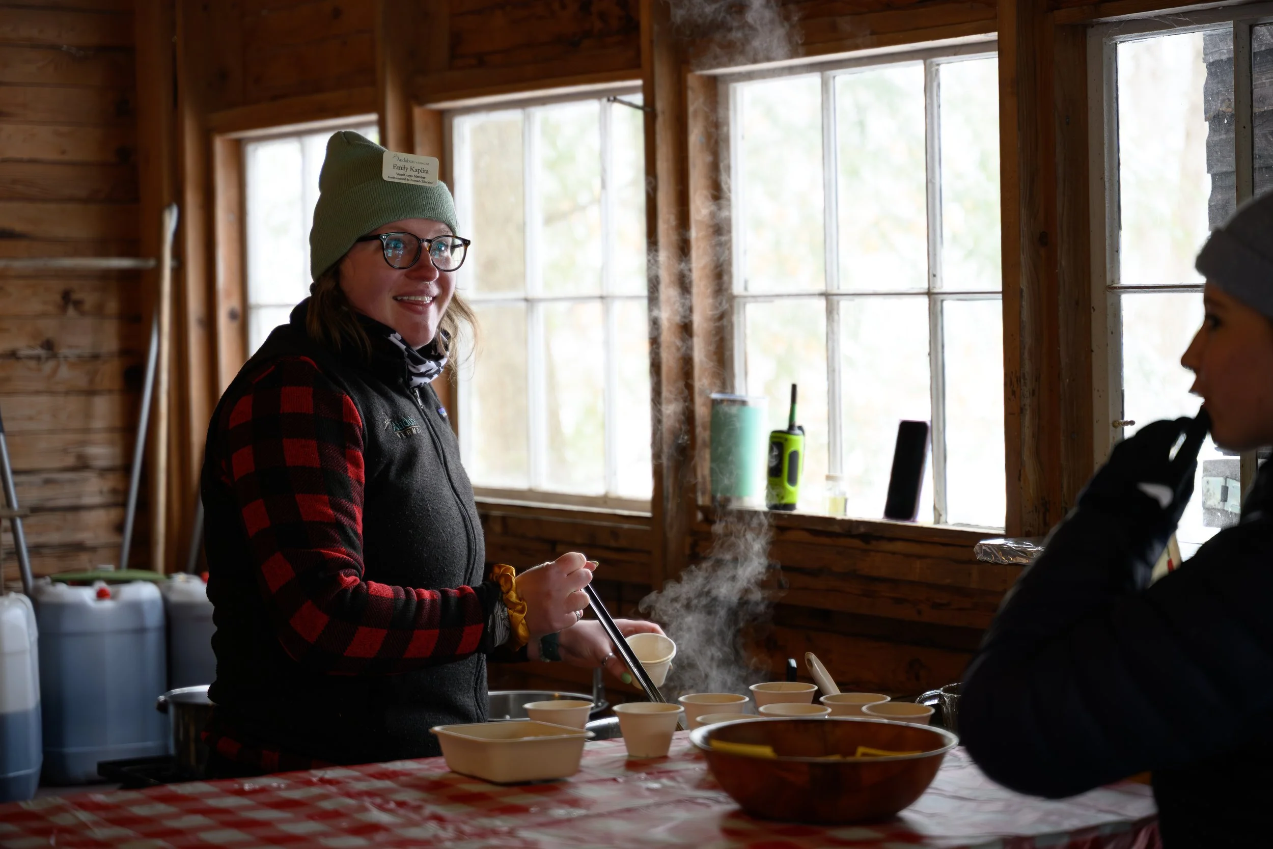 A woman wearing glasses, a green beanie, and a black vest with a red and black plaid shirt underneath, preparing food in a rustic wooden kitchen with large windows, steam rising from bowls on a checkered tablecloth, and another person partially visib