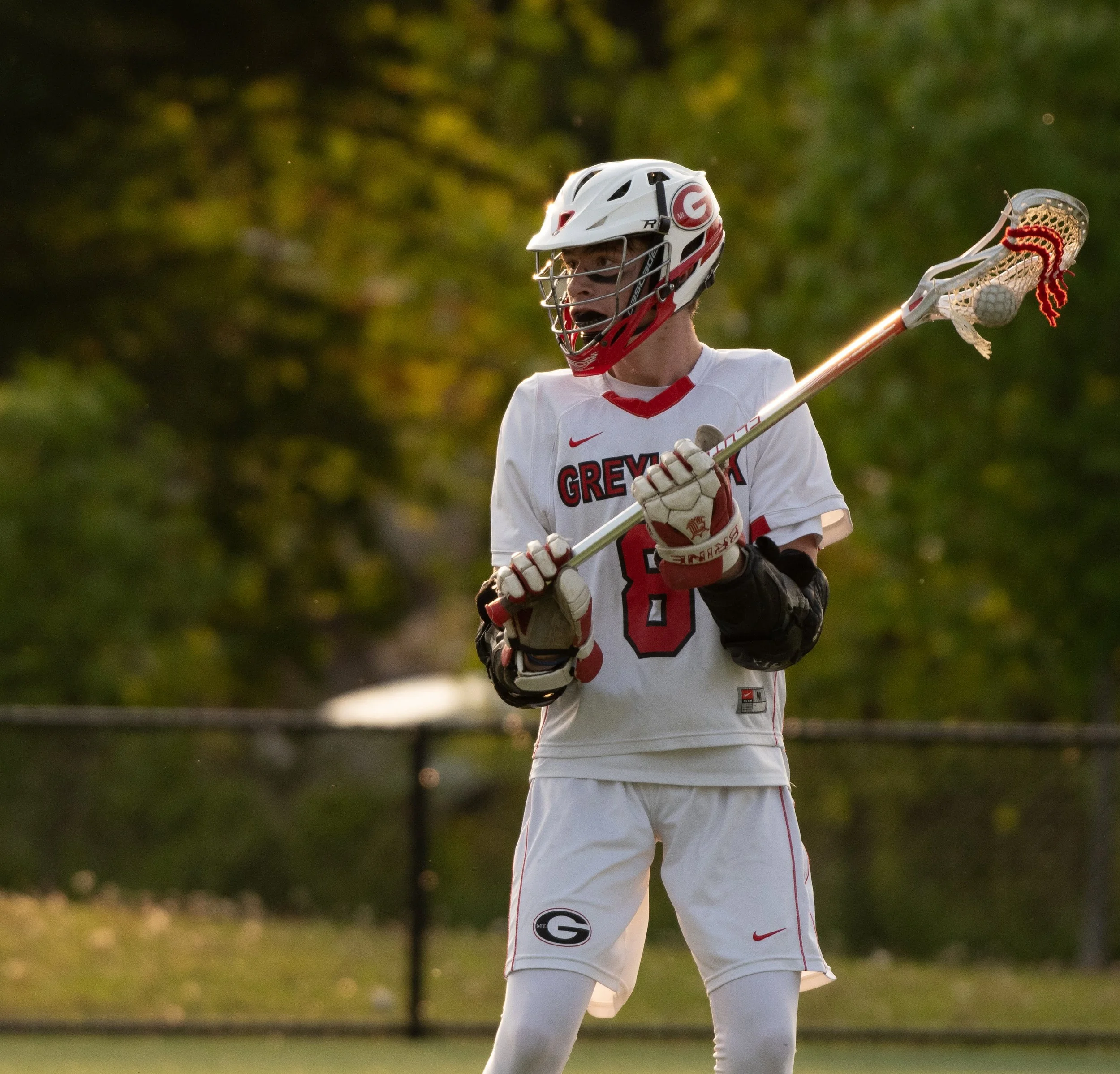 High school lacrosse player wearing a white uniform with "Greylock" logo and number 8, holding a lacrosse stick on a field.
