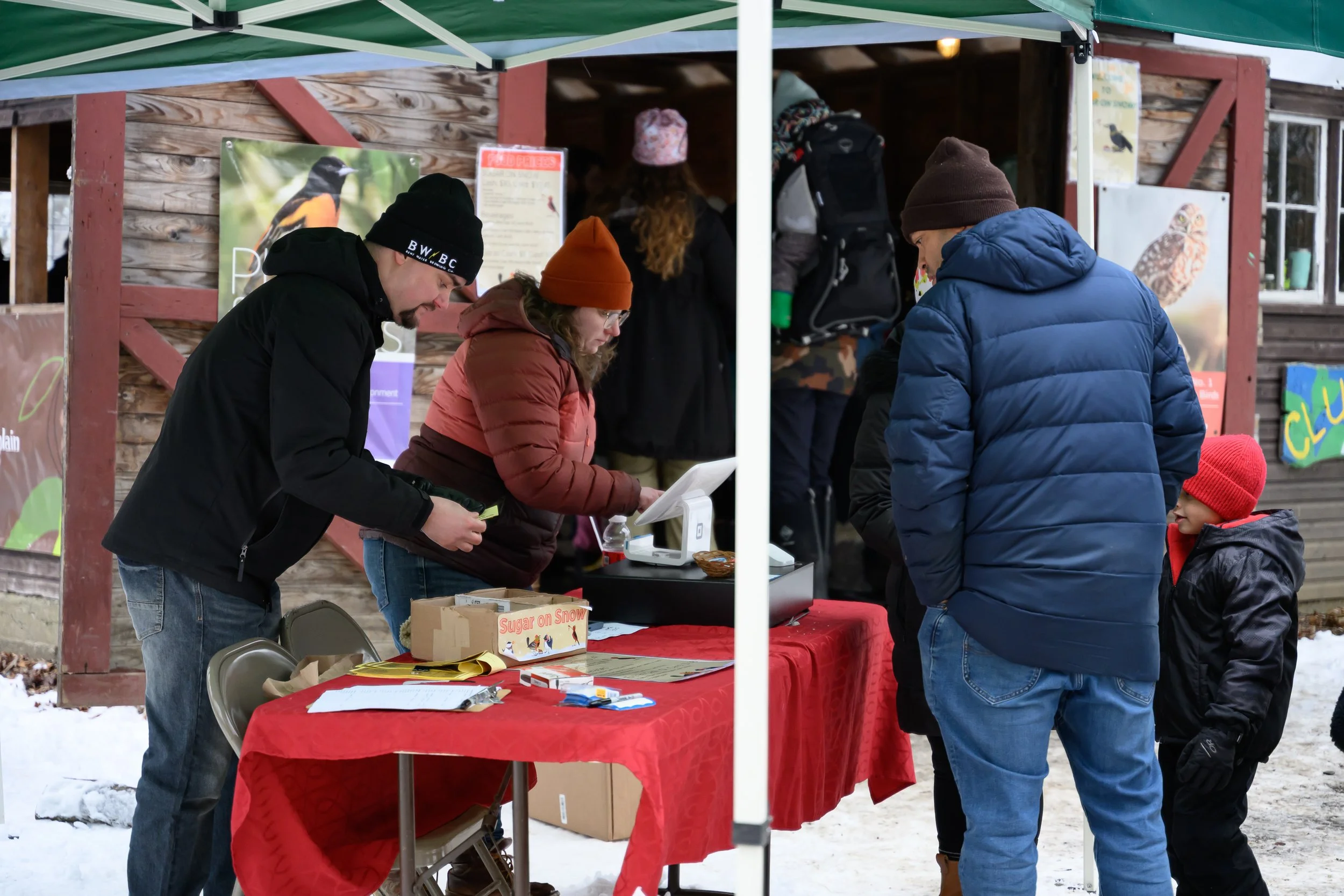 People attending an outdoor event at a booth with a red tablecloth, under a green canopy, in a snowy setting. They are dressed in winter clothing and appear to be engaging in a transaction or conversation.